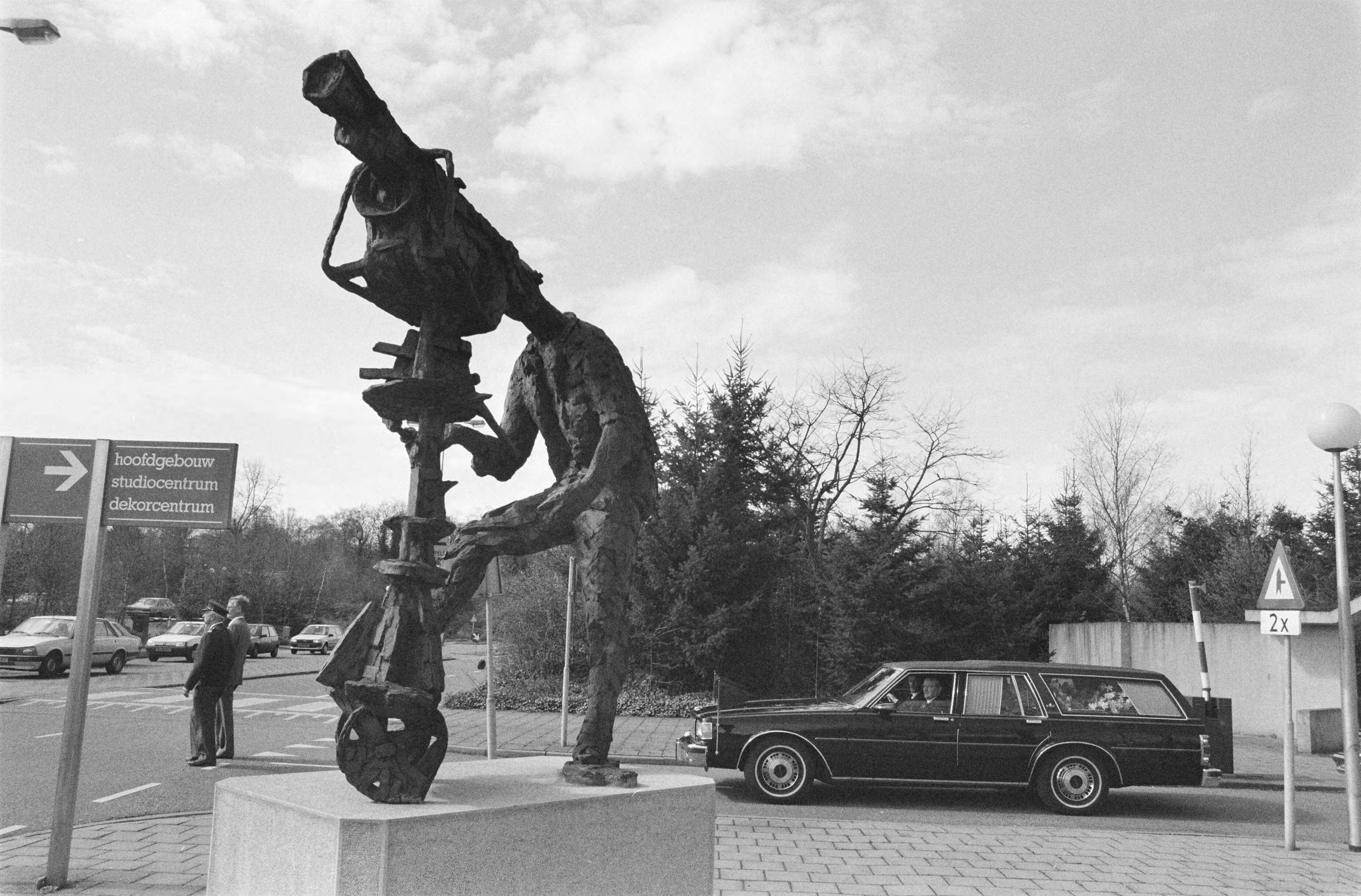 Herdenkingsdienst voor de doodgeschoten cameraman Cornel Lagrouw in Hilversum — De begrafenisstoet rijdt langs het beeld "de cameraman" op het NOS-terrein — Foto: Bogaerts, Rob / Anefo. Licentie: Set B: Rechtenvrij / Publiek Domein. Bron: Nationaal Archief. http://hdl.handle.net/10648/ad71d404-d0b4-102d-bcf8-003048976d84