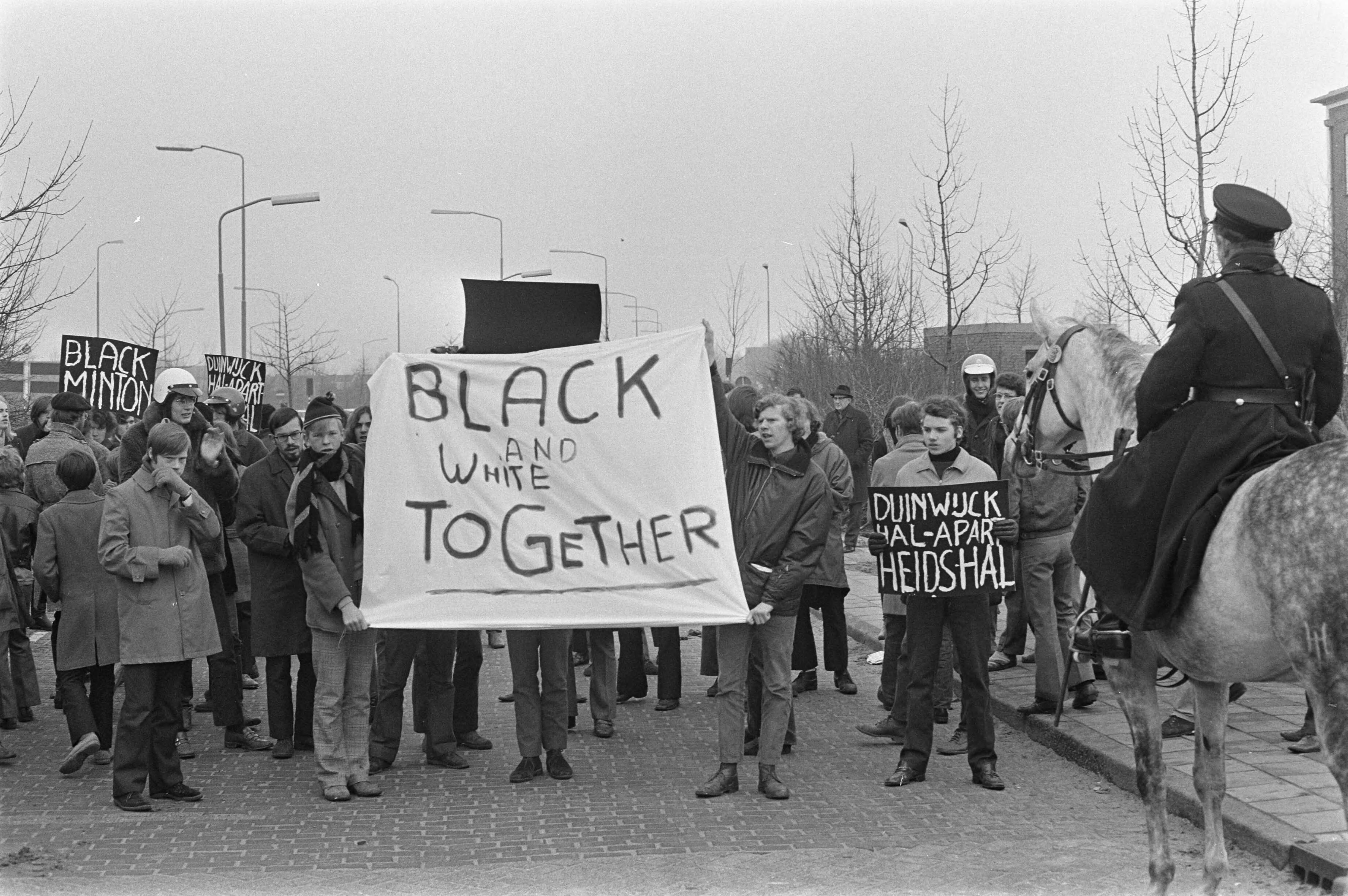Fotocollectie Anefo — Demonstranten met spandoeken tegen deelname van Zuid-Afrikaanse spelers bij Duinwijckhal Haarlem. 18 Demonstranten met borden, 19 Demonstrant op motorkap — Foto: Evers, Joost / Anefo. Licentie: Set B: Rechtenvrij / Publiek Domein. Bron: Nationaal Archief. http://hdl.handle.net/10648/ab8d4b50-d0b4-102d-bcf8-003048976d84