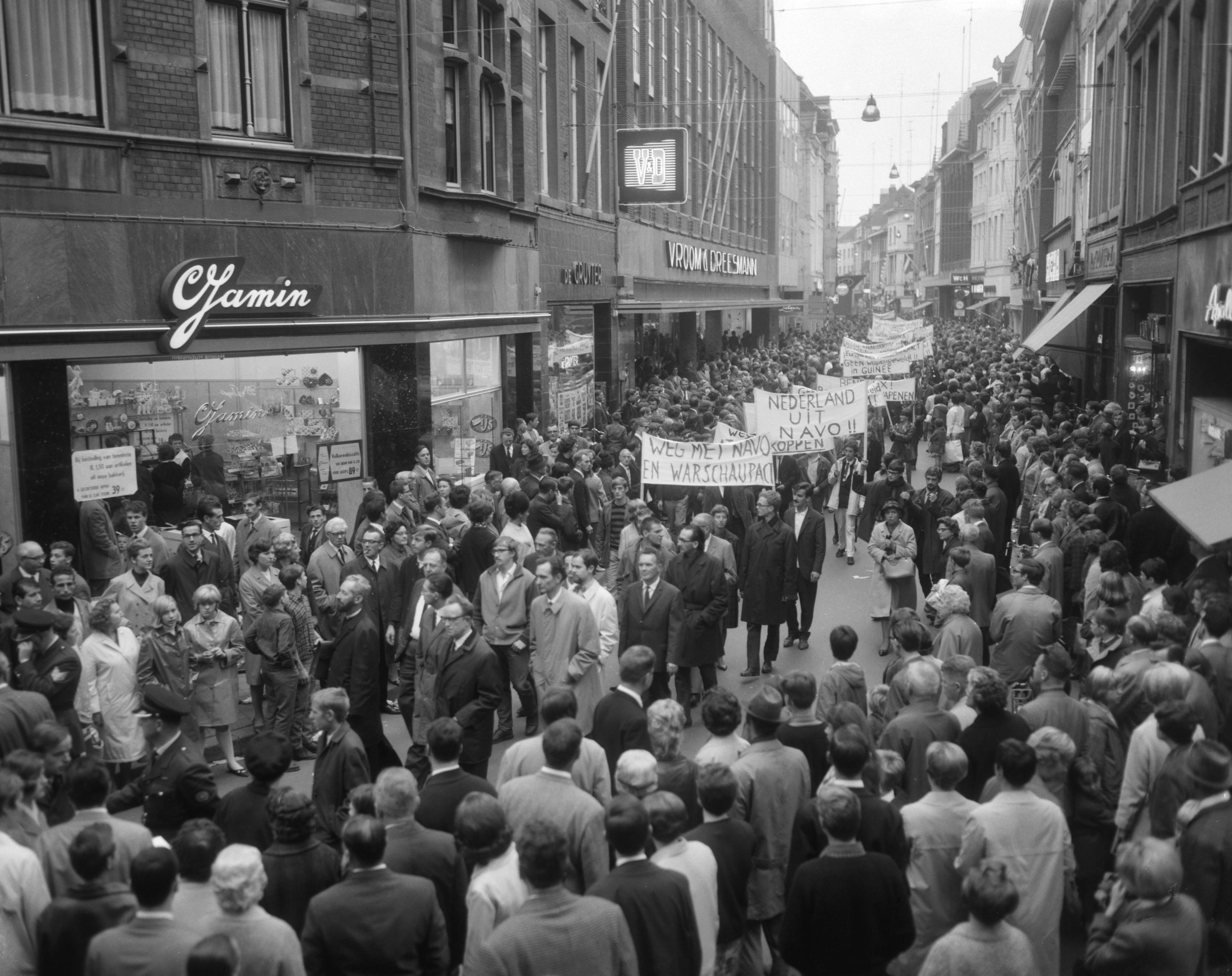 Anti NAVO-demonstratie in Maastricht — Demonstranten met spandoeken in de Grote Straat — Foto: Bilsen, Joop van / Anefo. Licentie: Set B: Rechtenvrij / Publiek Domein. Bron: Nationaal Archief. http://hdl.handle.net/10648/aafde1fe-d0b4-102d-bcf8-003048976d84