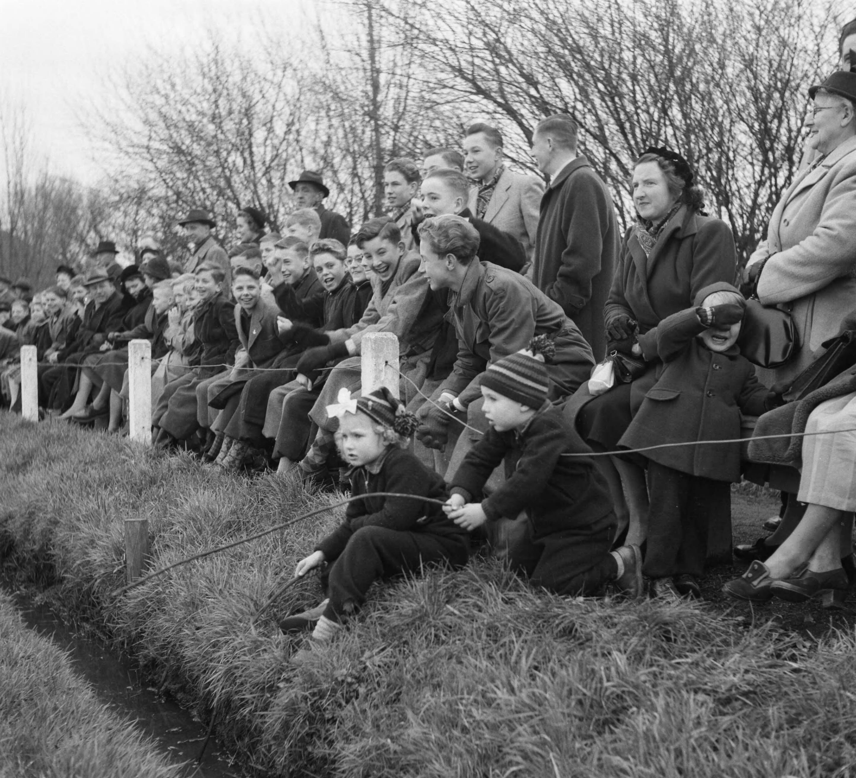Fotocollectie Anefo — Korfbal. Koog Zaandijk tegen Westerkwartier 4-7. Supporters langs het veld lachen. In de voorgrond twee kinderen die "vissen" in een greppel. Een kind rechts huilt — Foto: Noske, J.D. / Anefo. Licentie: Set B: Rechtenvrij / Publiek Domein. Bron: Nationaal Archief. http://hdl.handle.net/10648/a8f42a4e-d0b4-102d-bcf8-003048976d84