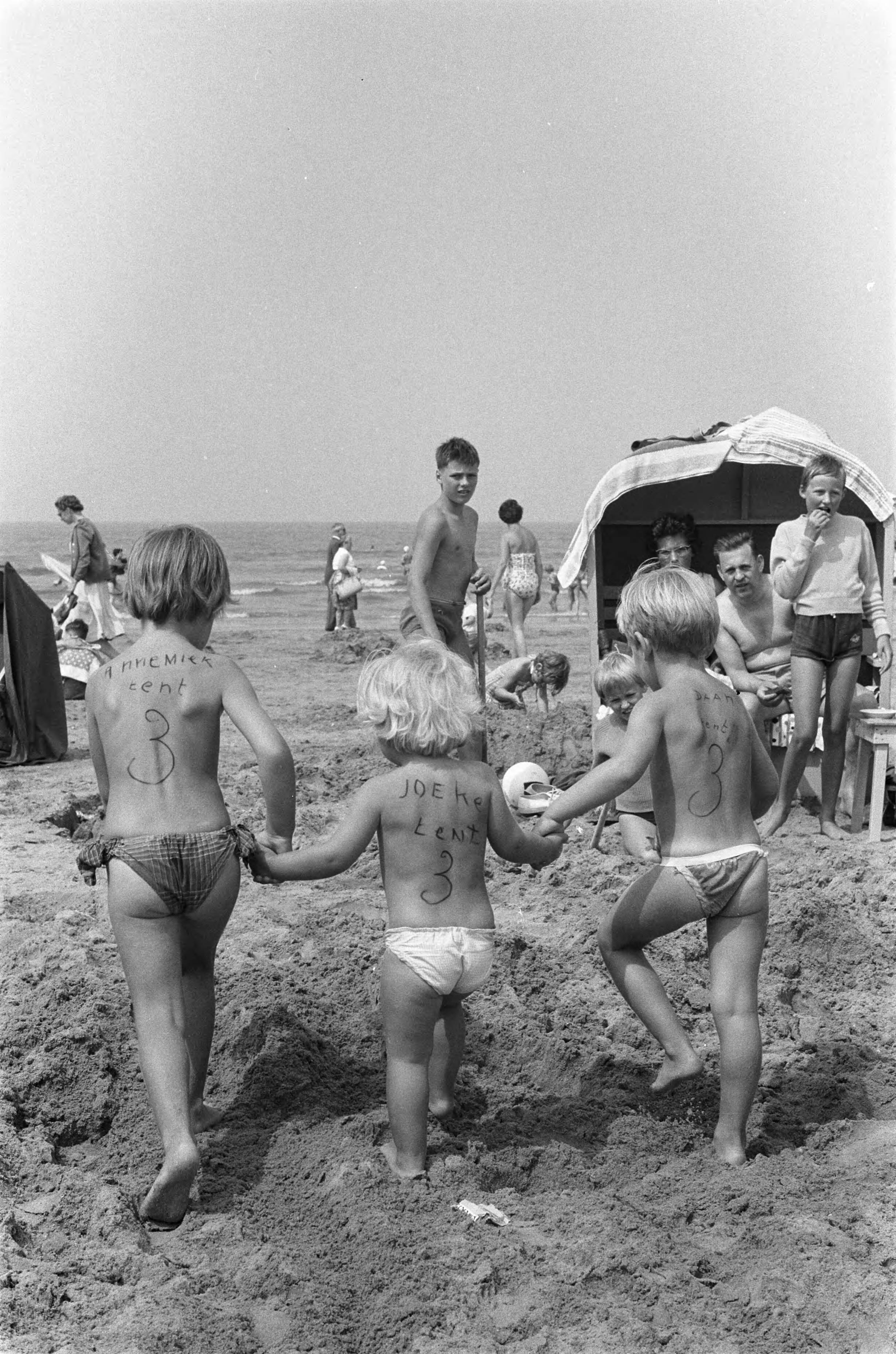 Fotocollectie Anefo — Zandvoort. Kinderen van dames Pot hebben namen op de rug geschreven en lopen hand in hand op het strand — Foto: Pot, Harry / Anefo. Licentie: Set B: Rechtenvrij / Publiek Domein. Bron: Nationaal Archief. http://hdl.handle.net/10648/a9de3058-d0b4-102d-bcf8-003048976d84