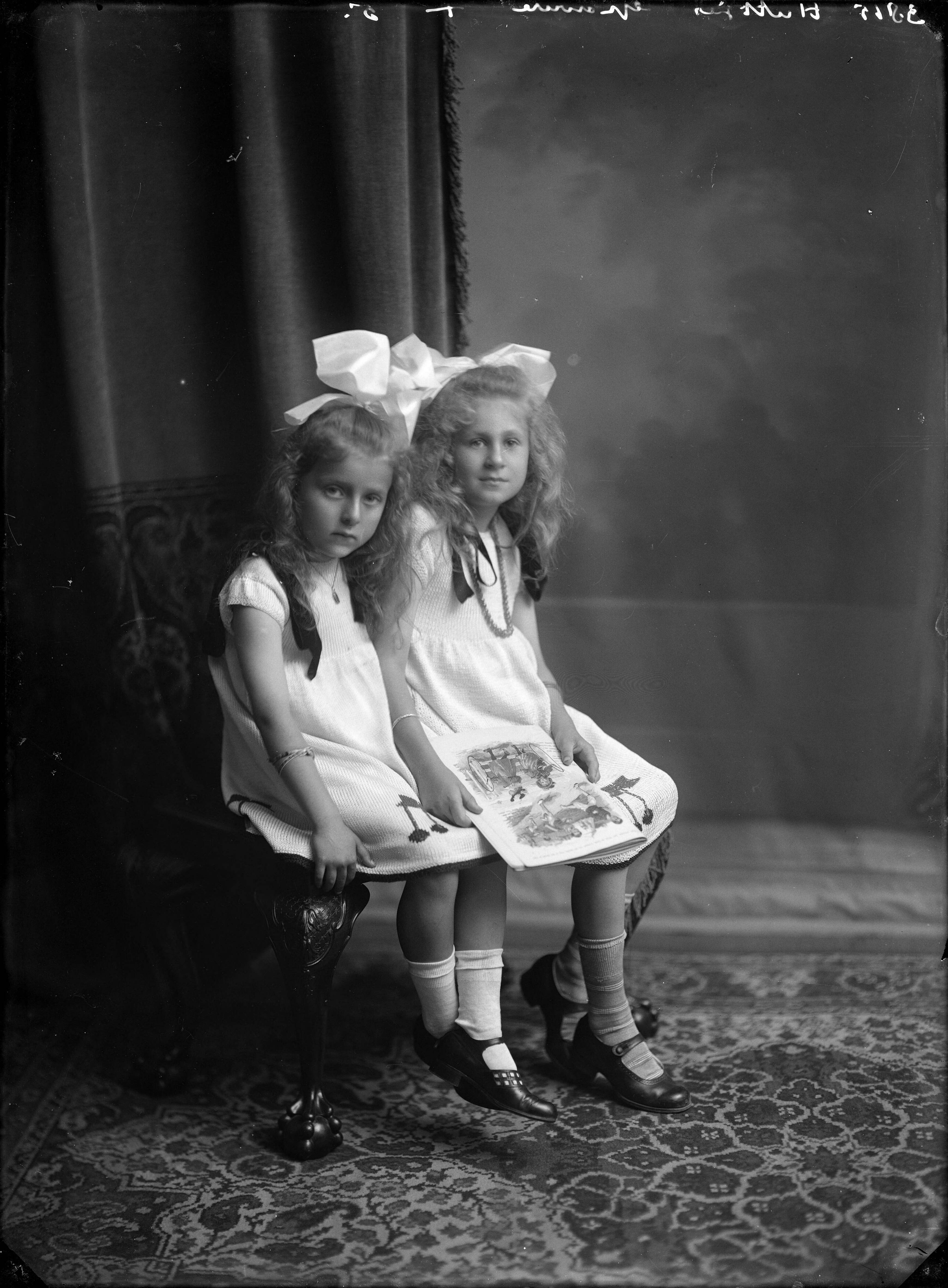 Portretten — Studio-opnames. Portret van twee meisjes op een bankje met lang haar, witte jurkjes, grote strikken en een boek op schoot. Haarlem, Nederland. 1918-1930. 3815 Huttjes gravure 5. — Foto: Berg, Th.C.J. van den / Spaarnestad Photo (05887). Licentie: Set B: Rechtenvrij / Publiek Domein. Bron: Nationaal Archief. http://hdl.handle.net/10648/35429da7-c7f5-a382-e063-6df0900a6686
