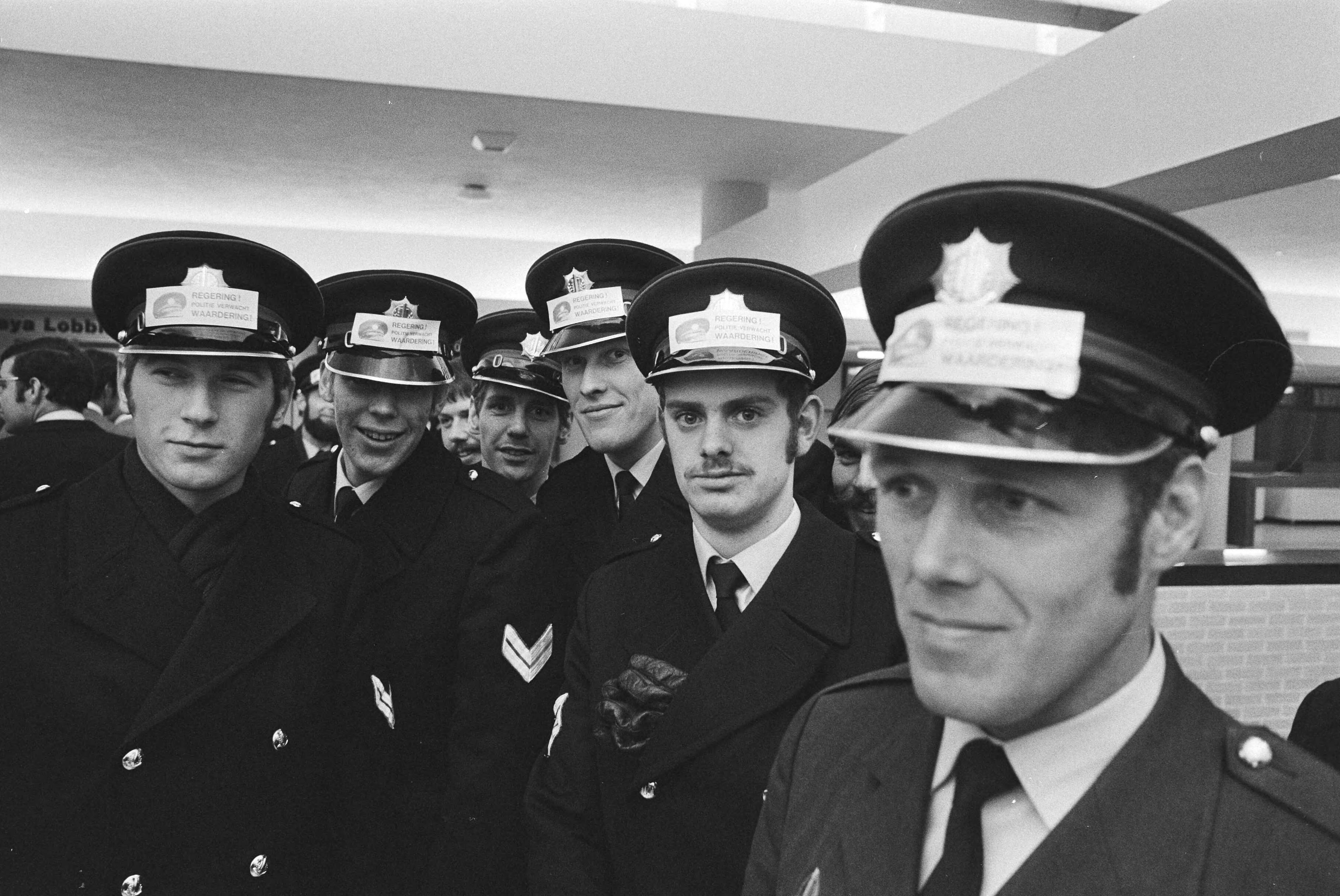 Fotocollectie Anefo — Protestvergadering van 2.000 politiemannen in Congresgebouw Den Haag ; o.a. voor betere salariering agenten met stickers op pet — Foto: Verhoeff, Bert / Anefo. Licentie: Set B: Rechtenvrij / Publiek Domein. Bron: Nationaal Archief. http://hdl.handle.net/10648/abac5680-d0b4-102d-bcf8-003048976d84