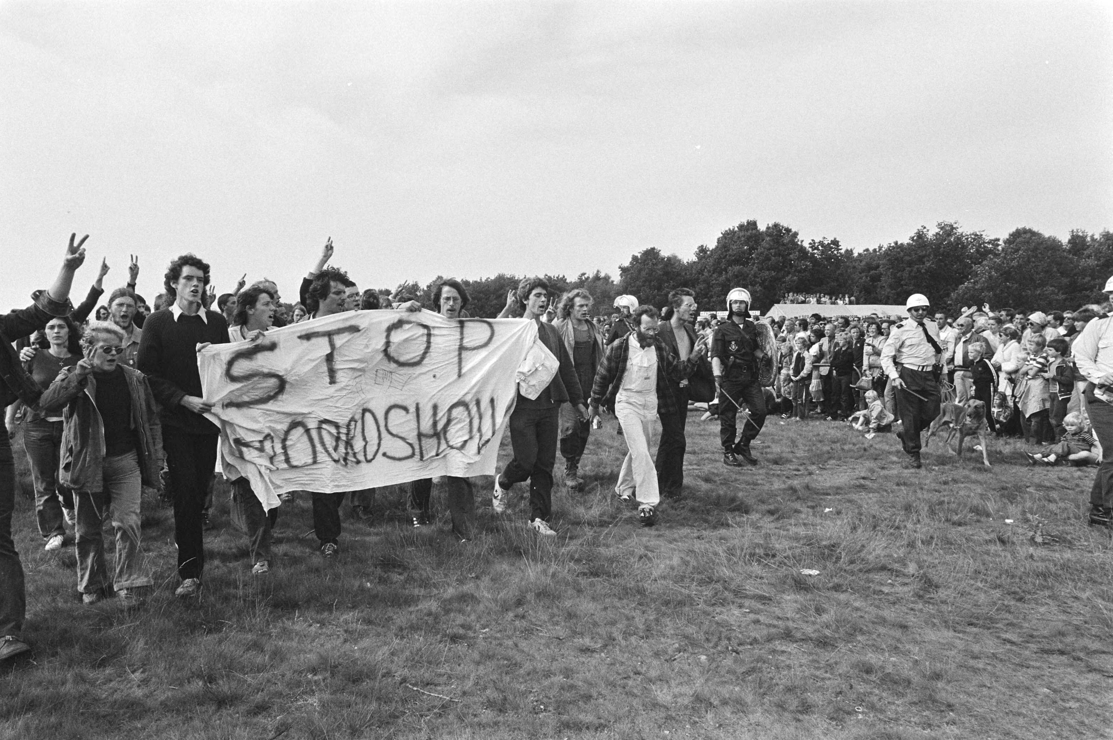 Open dag op luchtmachtbasis Soesterberg — Demonstranten van de actiegroep 'Onkruit' met spandoek 'Stop Moordshow' op de open dag op luchtmachtbasis Soesterberg terwijl de marechaussee een oogje in het zeil houdt — Foto: Dijk, Hans van / Anefo. Licentie: Set B: Rechtenvrij / Publiek Domein. Bron: Nationaal Archief. http://hdl.handle.net/10648/ad039af2-d0b4-102d-bcf8-003048976d84