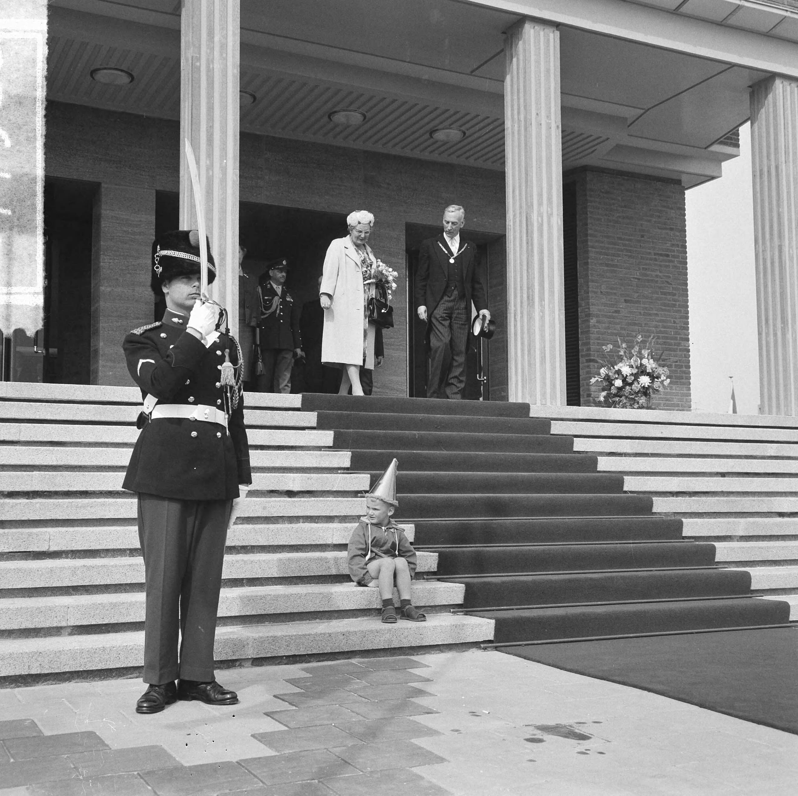 Koningin Juliana opent het nieuwe stadhuis te Vlissingen — Koningin Juliana verlaat het stadhuis, met naast zich burgemeester B. Kolff. Op de trappen zit een jongetje met feestmuts, recht brengt een marechaussee een saluut met zijn sabel — Foto: Nijs, Jac. de / Anefo. Licentie: Set B: Rechtenvrij / Publiek Domein. Bron: Nationaal Archief. http://hdl.handle.net/10648/bb9a8e3a-3ed9-fd13-7ac7-9b99dca8b160