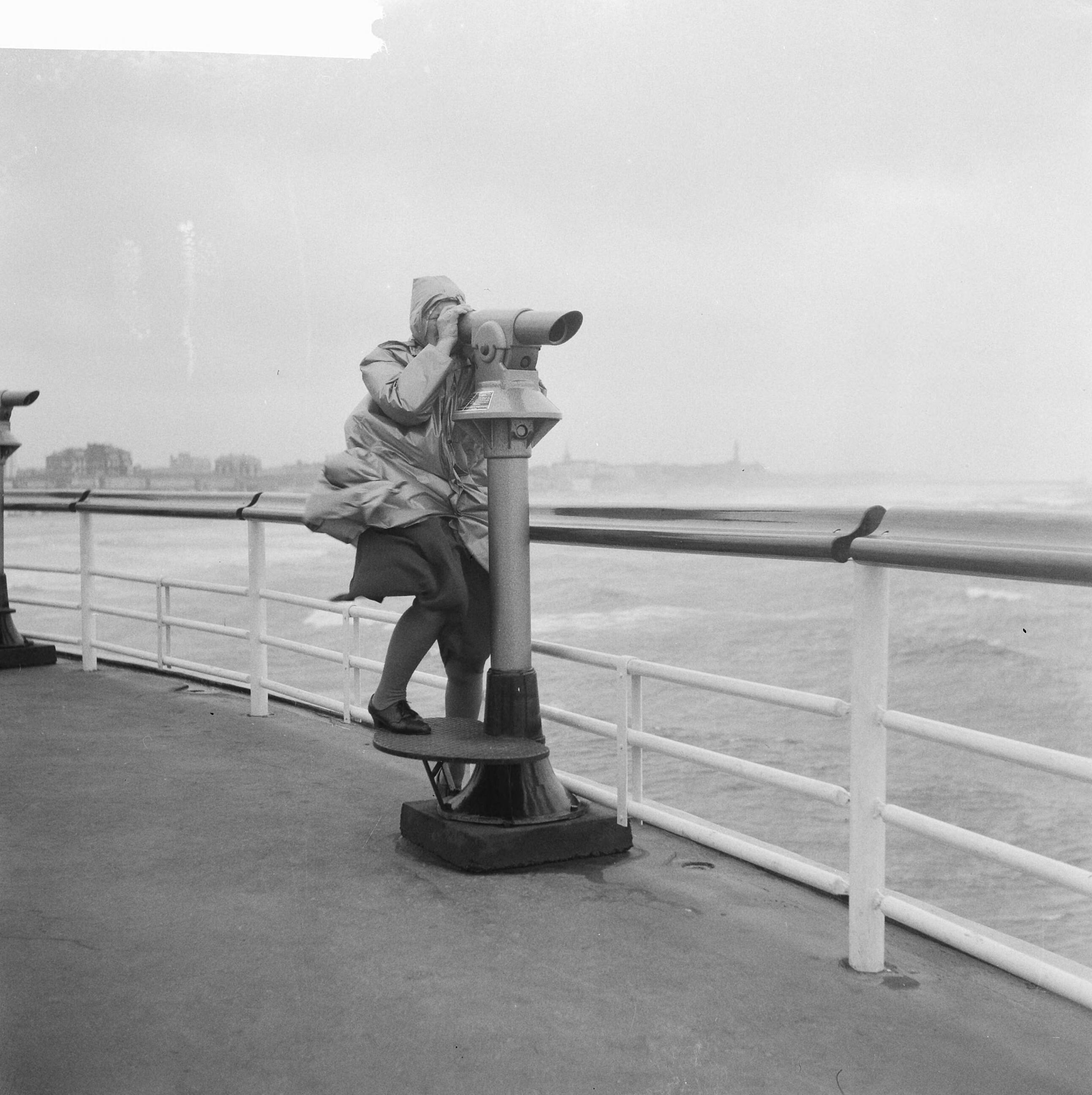 Fotocollectie Anefo — Strand van Scheveningen tijdens slecht weer. Kijken door een verrekijker op de Pier van Scheveningen — Foto: Koch, Eric / Anefo. Licentie: Set B: Rechtenvrij / Publiek Domein. Bron: Nationaal Archief. http://hdl.handle.net/10648/a9e02b7e-d0b4-102d-bcf8-003048976d84