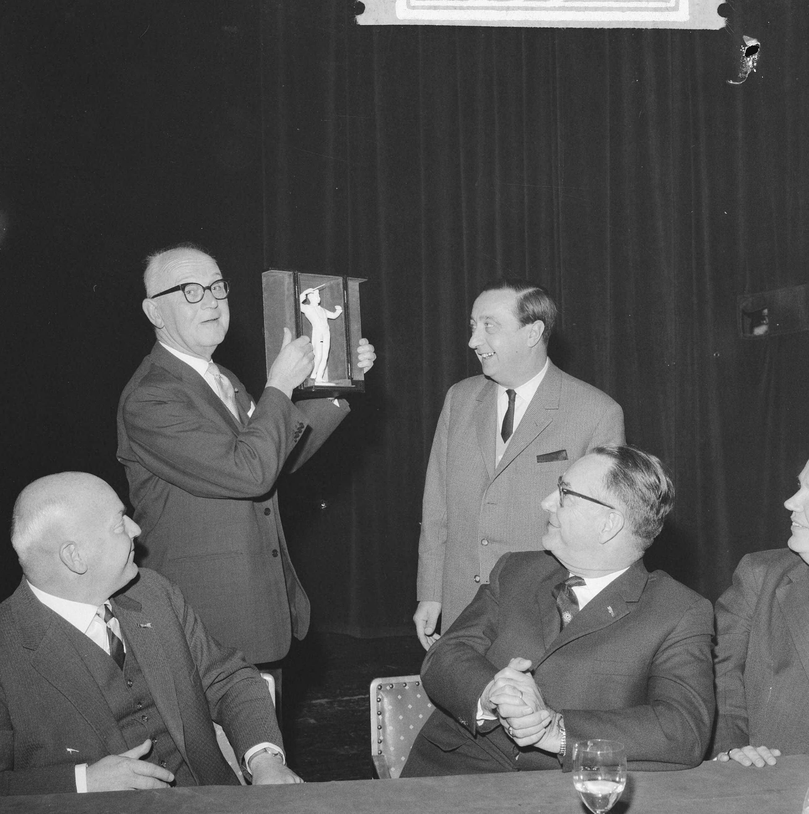 Fotocollectie Anefo — Jubileumcongres 50 jarig bestaan Vereniging van Nederlandse Gemeenten. Burgemeester van Den Haag H.A.M.T. Kolfschoten toont een doos met daarin een beeldje van een dirigent — Foto: Koch, Eric / Anefo. Licentie: Set B: Rechtenvrij / Publiek Domein. Bron: Nationaal Archief. http://hdl.handle.net/10648/aa0b6460-d0b4-102d-bcf8-003048976d84