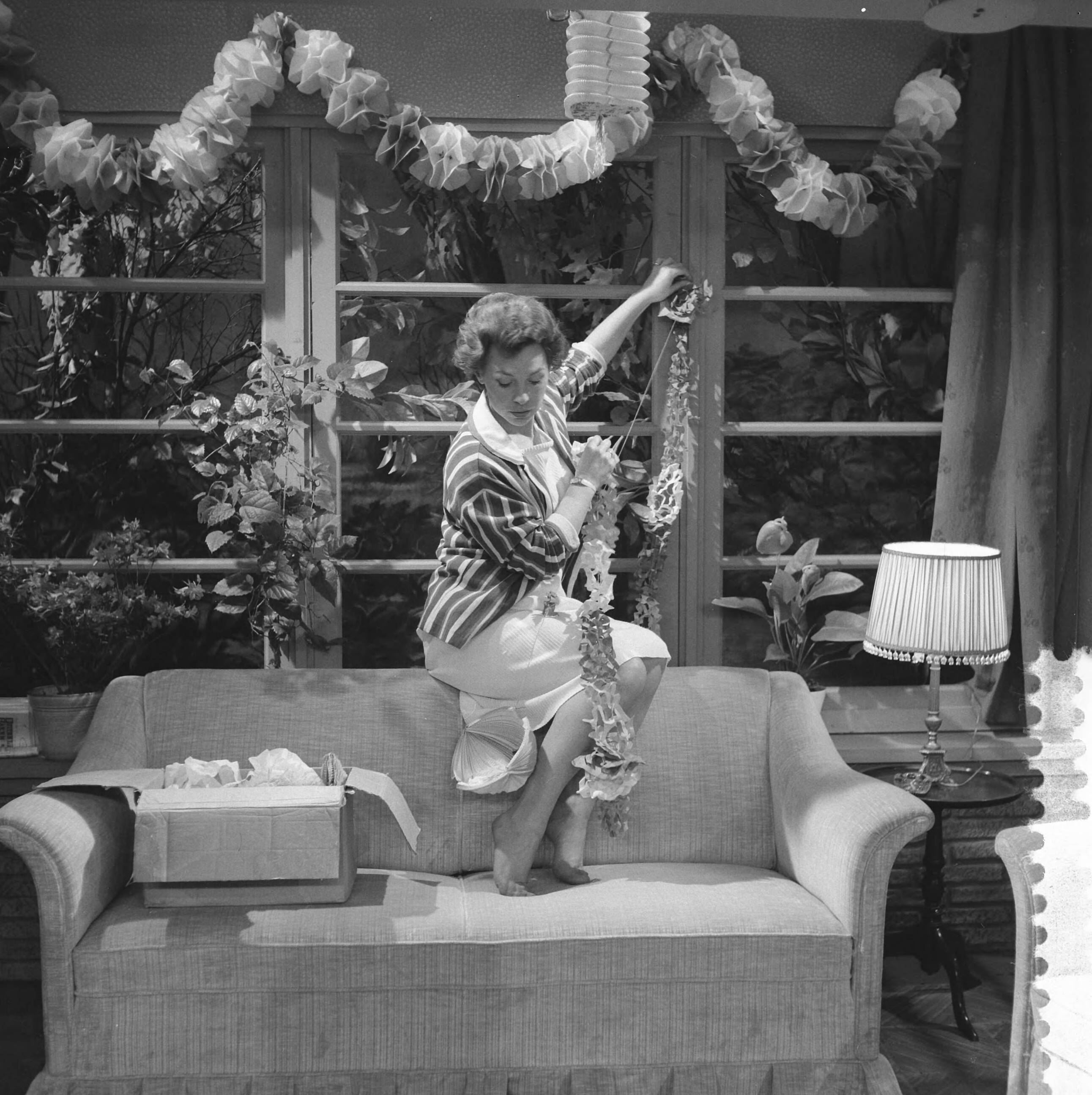Fotocollectie Anefo — VARA televisieserie "Bijna Twintig". Eenakter "Toen ik bijna twintig was¨van J.G. Toonder. Teddy Schaank (moeder) hangt slingers op — Foto: Lindeboom, Henk / Anefo. Licentie: Set B: Rechtenvrij / Publiek Domein. Bron: Nationaal Archief. http://hdl.handle.net/10648/a9cca158-d0b4-102d-bcf8-003048976d84