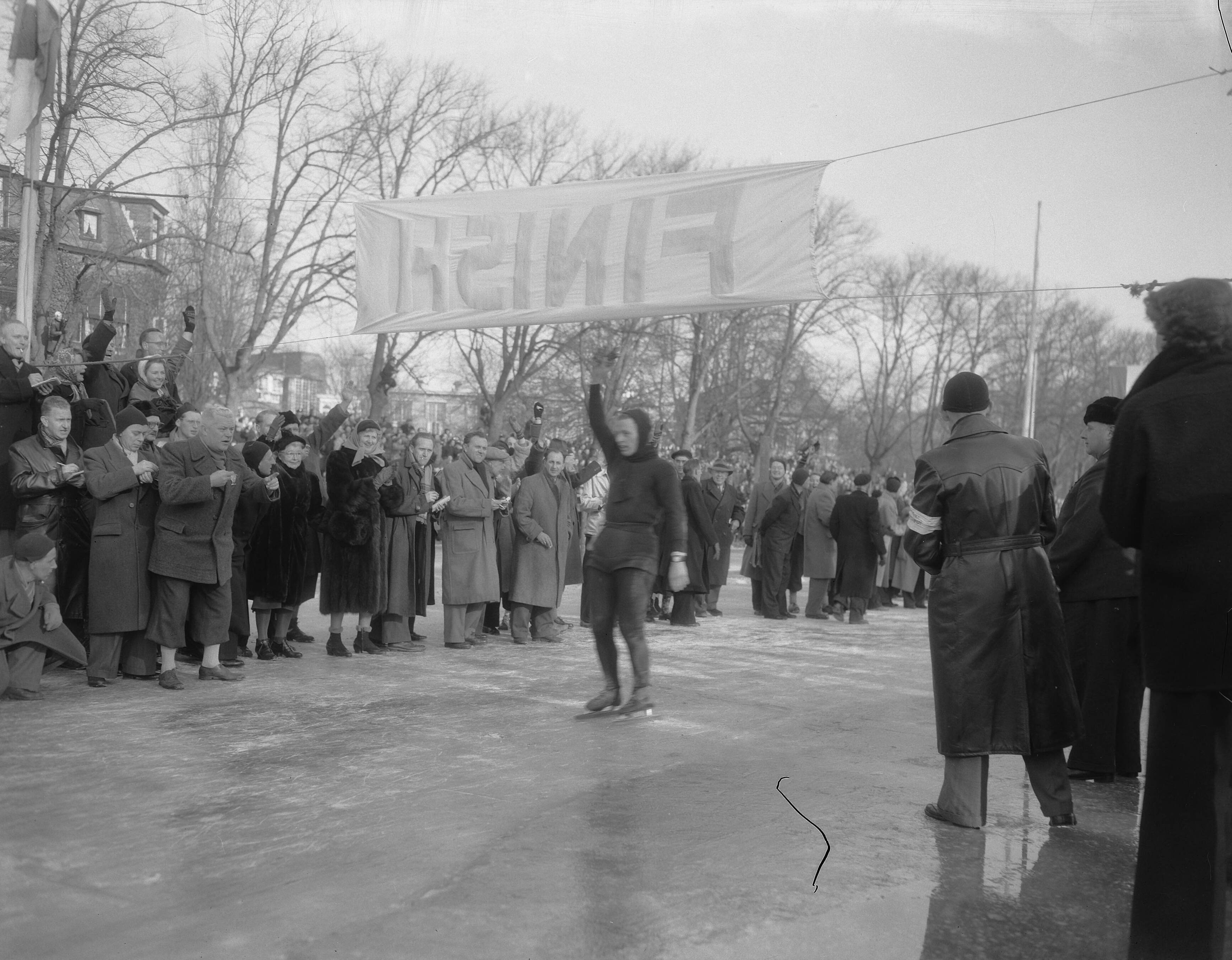 Elfstedentocht 1954 — Elfstedentocht. Jeen van den Berg gaat als eerste over de finish — Foto: Duinen, G.W. van / Anefo. Licentie: Set B: Rechtenvrij / Publiek Domein. Bron: Nationaal Archief. http://hdl.handle.net/10648/a91c681a-d0b4-102d-bcf8-003048976d84