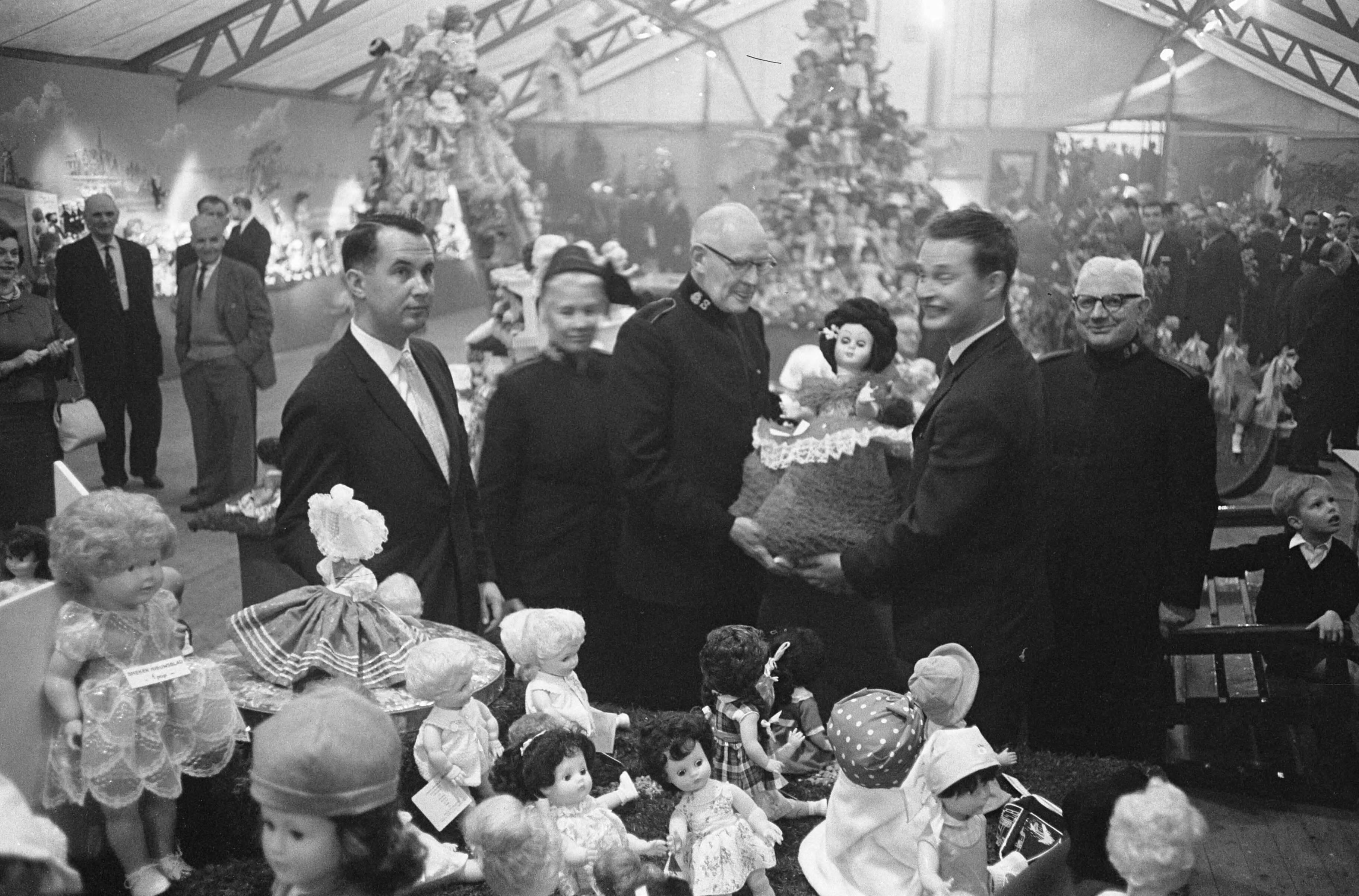 Fotocollectie Anefo — Albert Heijn N.V. in Zaandam heeft t.g.v. het 75-jarig bestaan aan haar vrienden en relaties gevraagd poppen te schenken i.p.v. de gebruikelijke cadeaus. Tijdens een receptie in Zaandam heeft de directie de poppen aan het Leger des Heils aangeboden. Links directeur J. Heijn, en rechts president-directeur A. Heijn die een pop overhandigt aan de landelijk commandant, W.F. Palstra — Foto: Pot, Harry / Anefo. Licentie: Set B: Rechtenvrij / Publiek Domein. Bron: Nationaal Archief. http://hdl.handle.net/10648/aa0a2604-d0b4-102d-bcf8-003048976d84