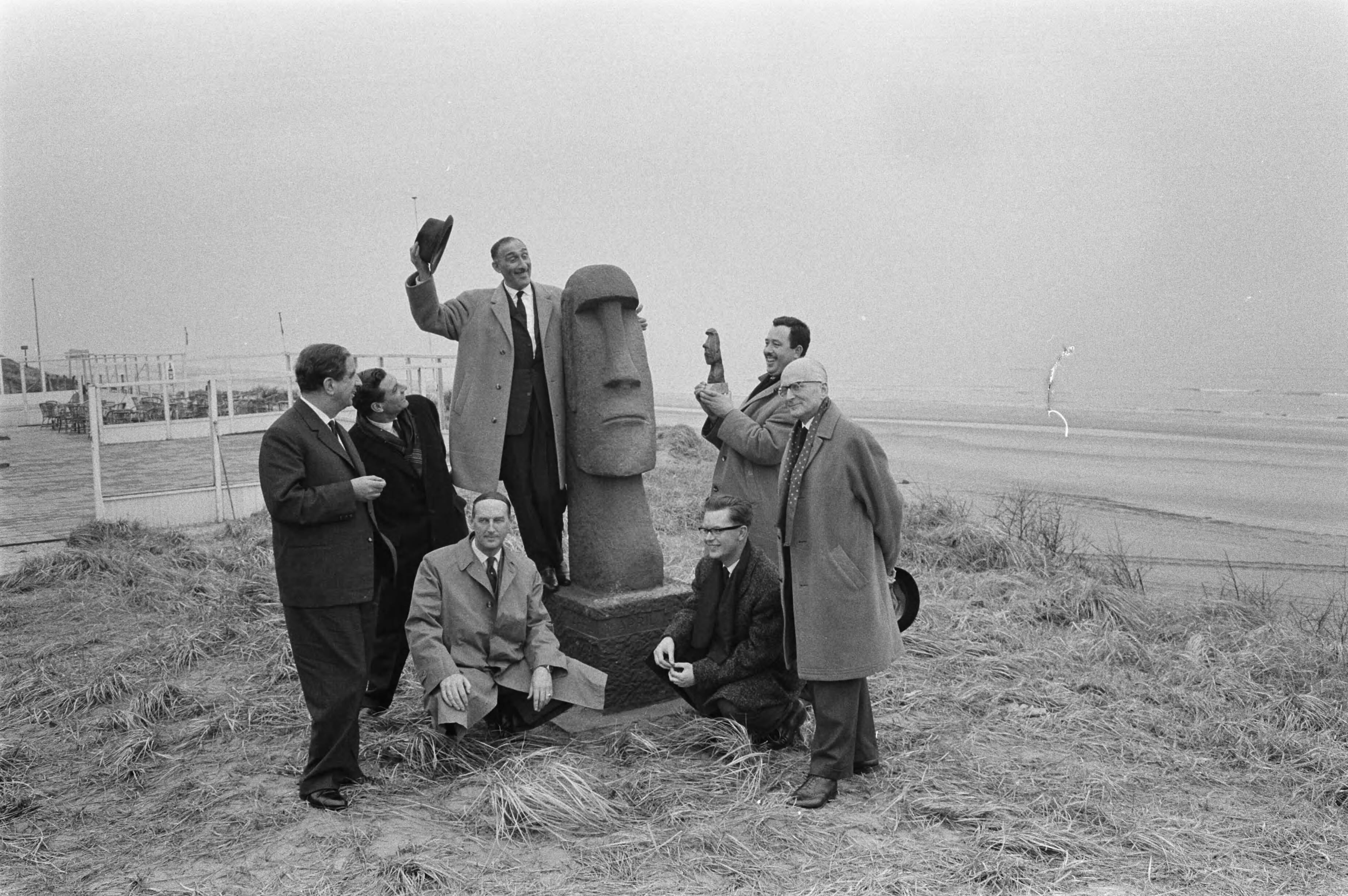 Fotocollectie Anefo — Nederland krijgt een „grapjas”-prijs, die jaarlijks op 1 april in Zandvoort zal worden uitgereikt voor de best geslaagde grap van het afgelopen jaar. De prijs bestaat uit een 31 cm hoog bronzen beeldje op marmeren voetstuk, een verkleind model van het verleden jaar op 1 april te Zandvoort „aangespoelde" beeld van het Paaseiland. Gisteren is te Zandvoort het uit 11 man bestaande „1-april-comité" geïnstalleerd. Hierboven een aantal leden van dit comité bij het beeld. V. I. n. r. staande- G. W. Oskamp, directeur provinciale VVV, B. Burmeister, directeur VVV-Delft; op het beeld Max Tailleur, Edo Tetterode, maker van het beeld en met verkleinde „grapjas"-prijs en burgemeester mr. H.M. van Fenema, voorzitter van het comité. Zittend J.B.Th. Hugenholtz, directeur van het Zandvoortse circuit en rechts G. F. Donner — Foto: Pot, Harry / Anefo. Licentie: Set B: Rechtenvrij / Publiek Domein. Bron: Nationaal Archief. http://hdl.handle.net/10648/aa2e6550-d0b4-102d-bcf8-003048976
