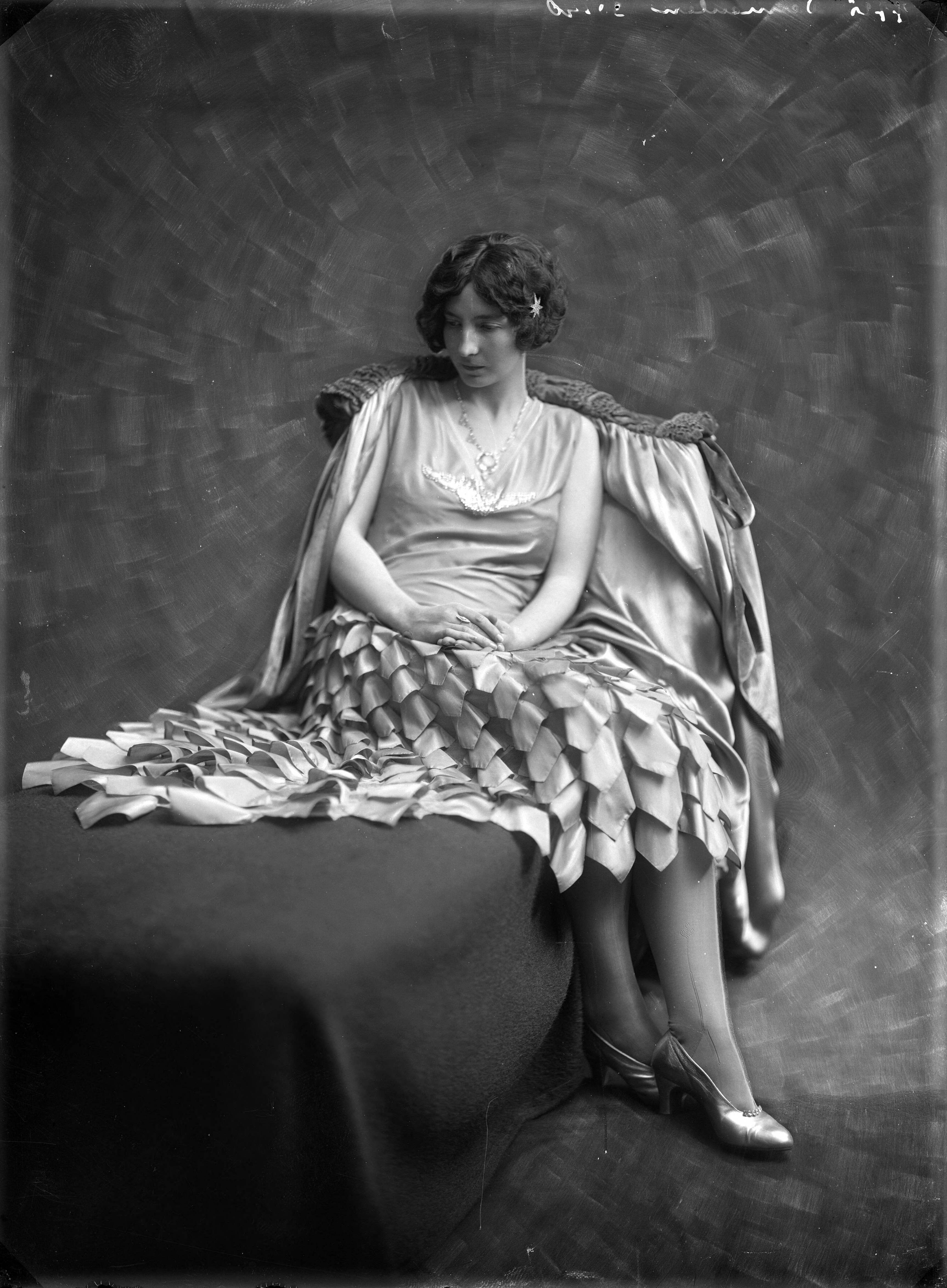 Mode, beauty — Studio-opname van een vrouw zittend op een bank in een maskeradejurk met puntstrookjes genaaid op de rok en met een fluwelen cape over de rugleuning. 7725 Vermeulen 30140. Haarlem, Nederland 1918-1930. — Foto: Berg, Th.C.J. van den / Spaarnestad Photo (05887). Licentie: Set B: Rechtenvrij / Publiek Domein. Bron: Nationaal Archief. http://hdl.handle.net/10648/35429da7-c550-a382-e063-6df0900a6686