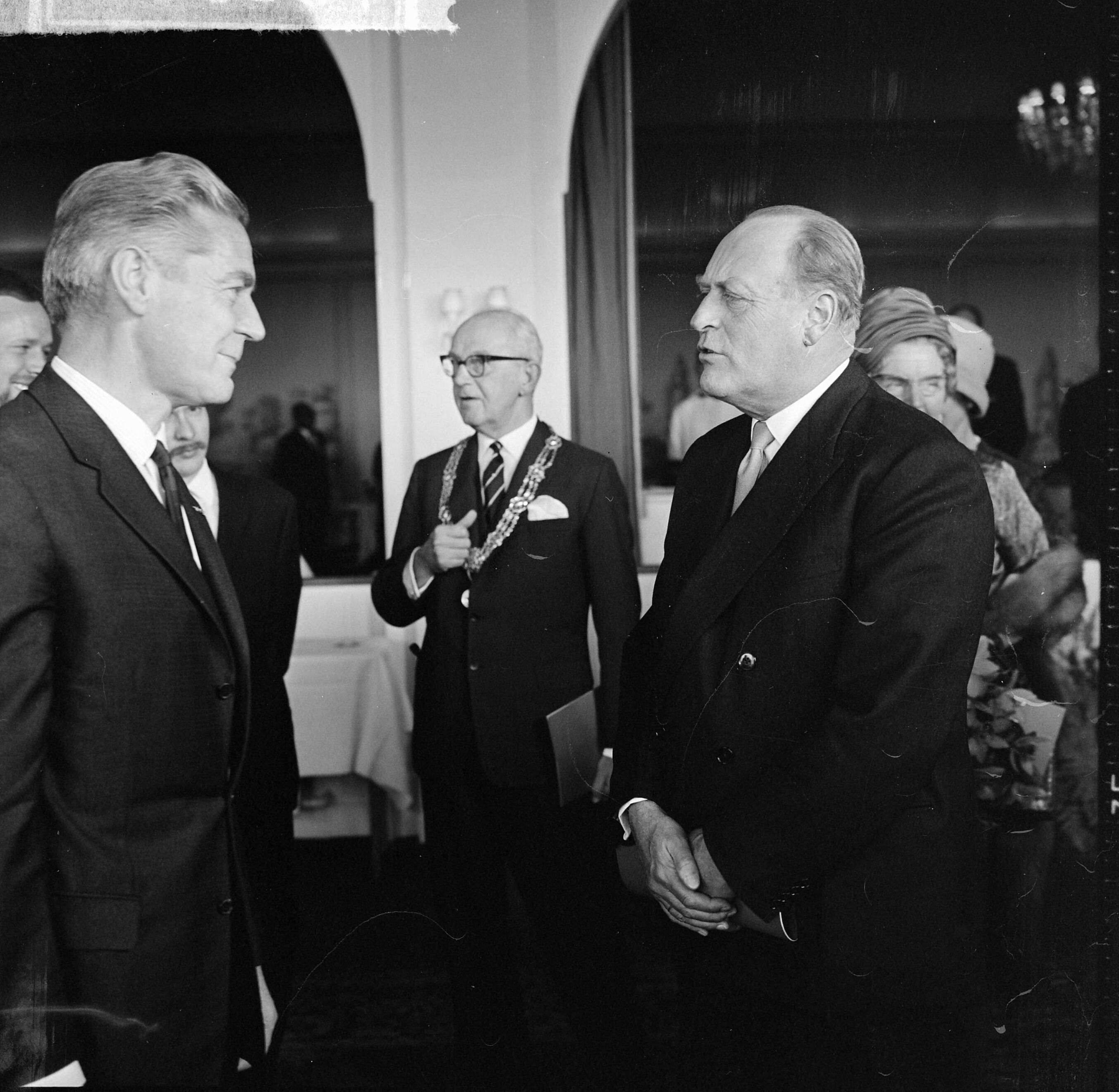 Staatsbezoek koning Olav van Noorwegen 1964 — Bezoek koning Olav van Noorwegen aan Den Haag. Koning Olav in gesprek met cineast Bert Haanstra. In het midden burgemeester Kolfschoten — Foto: Koch, Eric / Anefo. Licentie: Set B: Rechtenvrij / Publiek Domein. Bron: Nationaal Archief. http://hdl.handle.net/10648/aa787992-d0b4-102d-bcf8-003048976d84