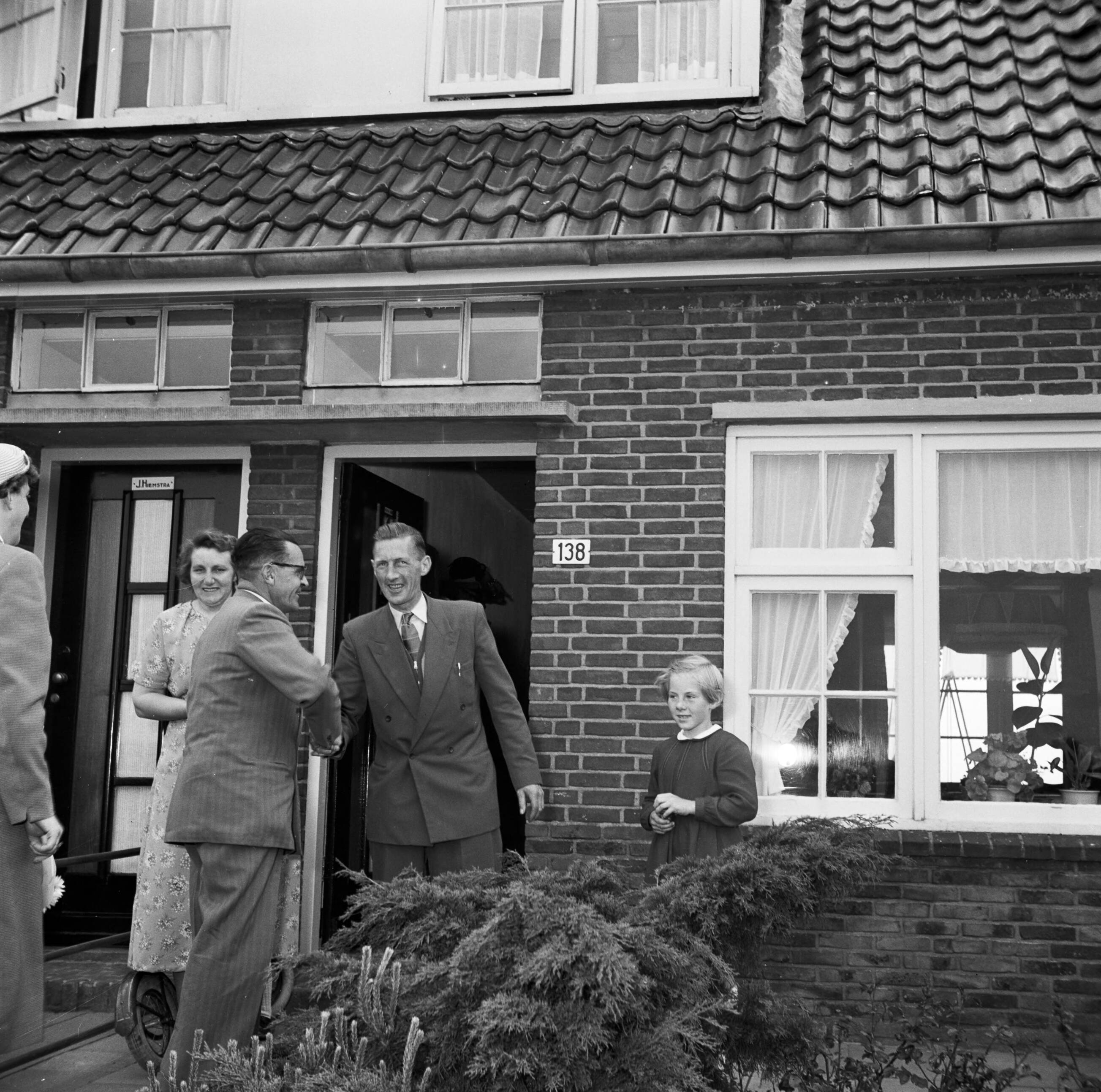 Fotocollectie Van de Poll — Gerard de Jong bij de voordeur van zijn woning, schudt de hand van Ynse Postma. Links zijn echtgenoten en rechts zijn dochterje Marietje — Foto: Poll, Willem van de. Licentie: Set B: Rechtenvrij / Publiek Domein. Bron: Nationaal Archief. http://hdl.handle.net/10648/aecce0b4-d0b4-102d-bcf8-003048976d84
