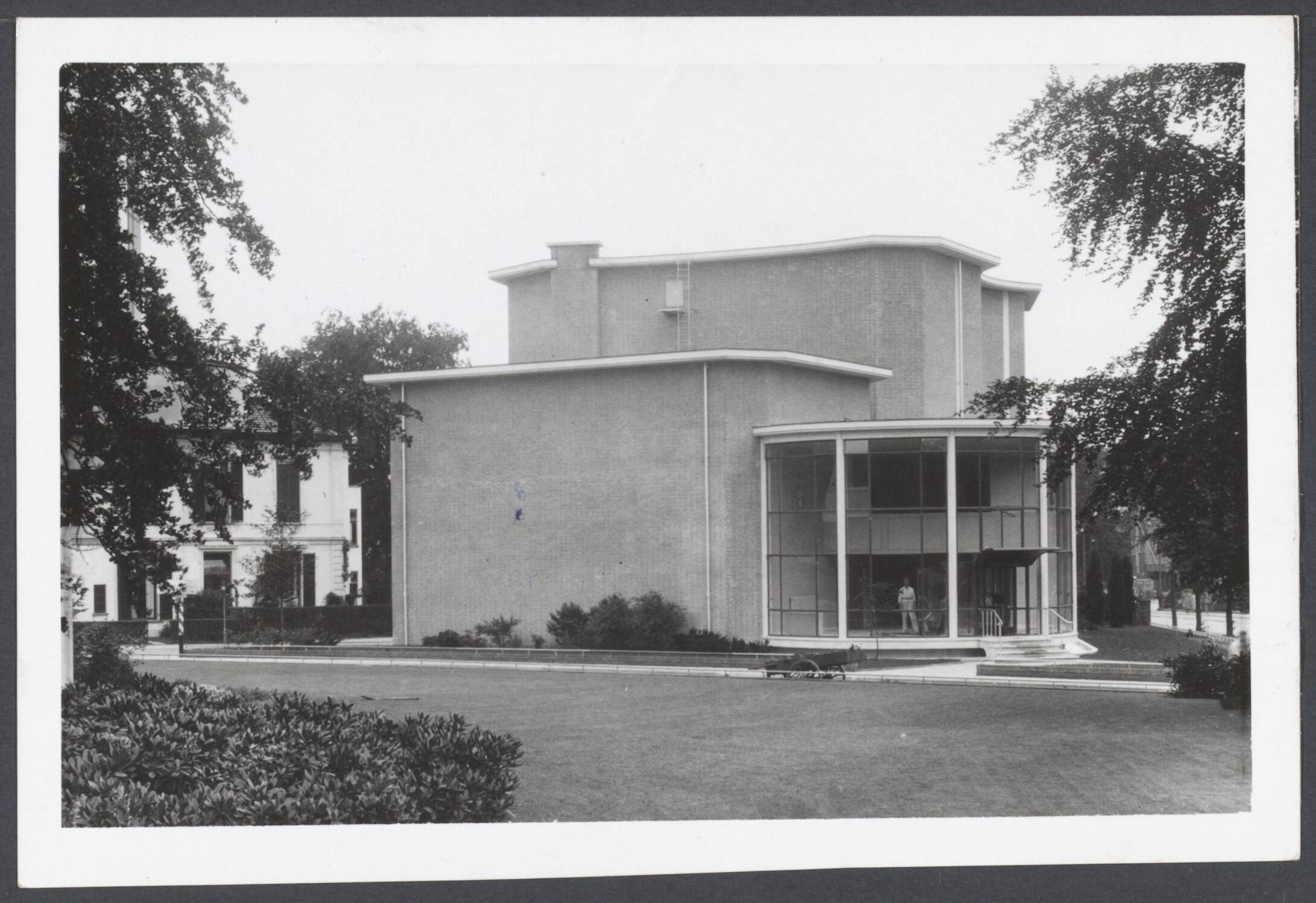 AVRO (Algemene Vereniging Radio Omroep). Studio-gebouwen, exterieur en interieur - Nederland — Exterieur van het in aanbouw zijnde nieuwe A.V.R.O. Studio te Hilversum, welke men over een maand in gebruik hoopt te kunnen nemen. Dit paviljoen sluit harmonisch geheel aan bij de reeds bestaande studio-gebouwen, de nieuwe grote studio-zaal heeft ongeveer de vorm van een vioolkast, waarin geen enkele rechte hoek te vinden is en waarvan de wanden niet evenwijdig lopen, waardoor het geluid zo snel mogelijk diffuus wordt, d.w.z. dat het zich verspreidt zonder dat vagebonderende geluidsstromen ontstaan — Foto: N.V. Polygoon. Licentie: Set B: Rechtenvrij / Publiek Domein. Bron: Nationaal Archief. http://hdl.handle.net/10648/798070b7-3c93-1fb3-8944-4acd81ef7b68