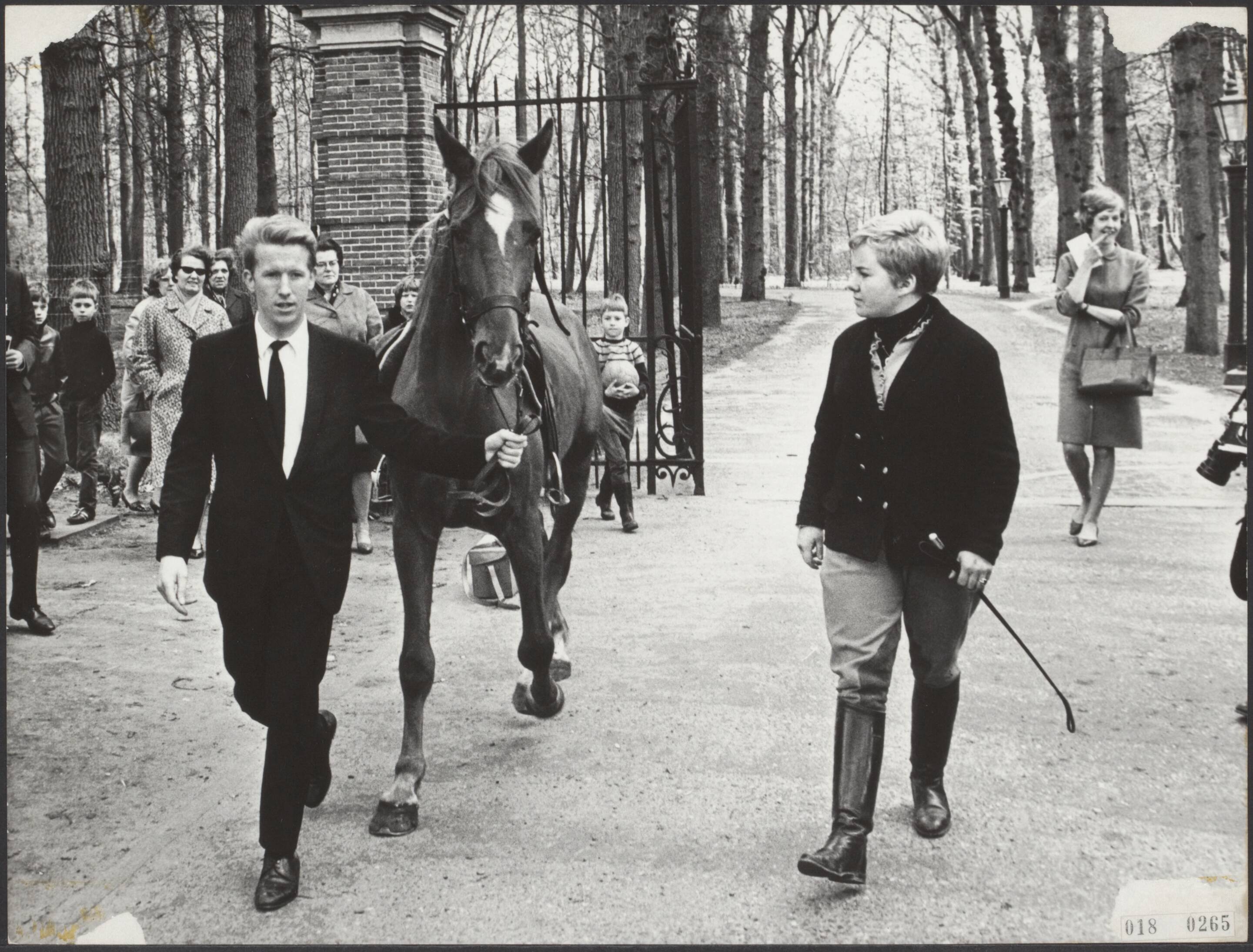 Fotocollectie Elsevier Binnenland — Even na het middaguur werd de paleiswacht van Drakensteyn opgeschrikt door een op hol geslagen paard, dat in volle ren de poort binnen draafde. Erop zat André Gorel, ober in hotel De Lage Vuursche, die het paard in de stal zou zetten, nadat mej. Diny Meewis er al moeilijkheden mee had. Nadat het paard door 8 marechaussees tot staan was gebracht in de paleistuin, kwam ober André Gorel terug naar de poort, waar mej. Diny Meewis stond te wachten, waarna zij samen het inmiddels gekalmeerde paard naar de stal terugbrachten — Foto: Koch, Eric / Anefo. Licentie: Set B: Rechtenvrij / Publiek Domein. Bron: Nationaal Archief. http://hdl.handle.net/10648/adc1bb7c-d0b4-102d-bcf8-003048976d84