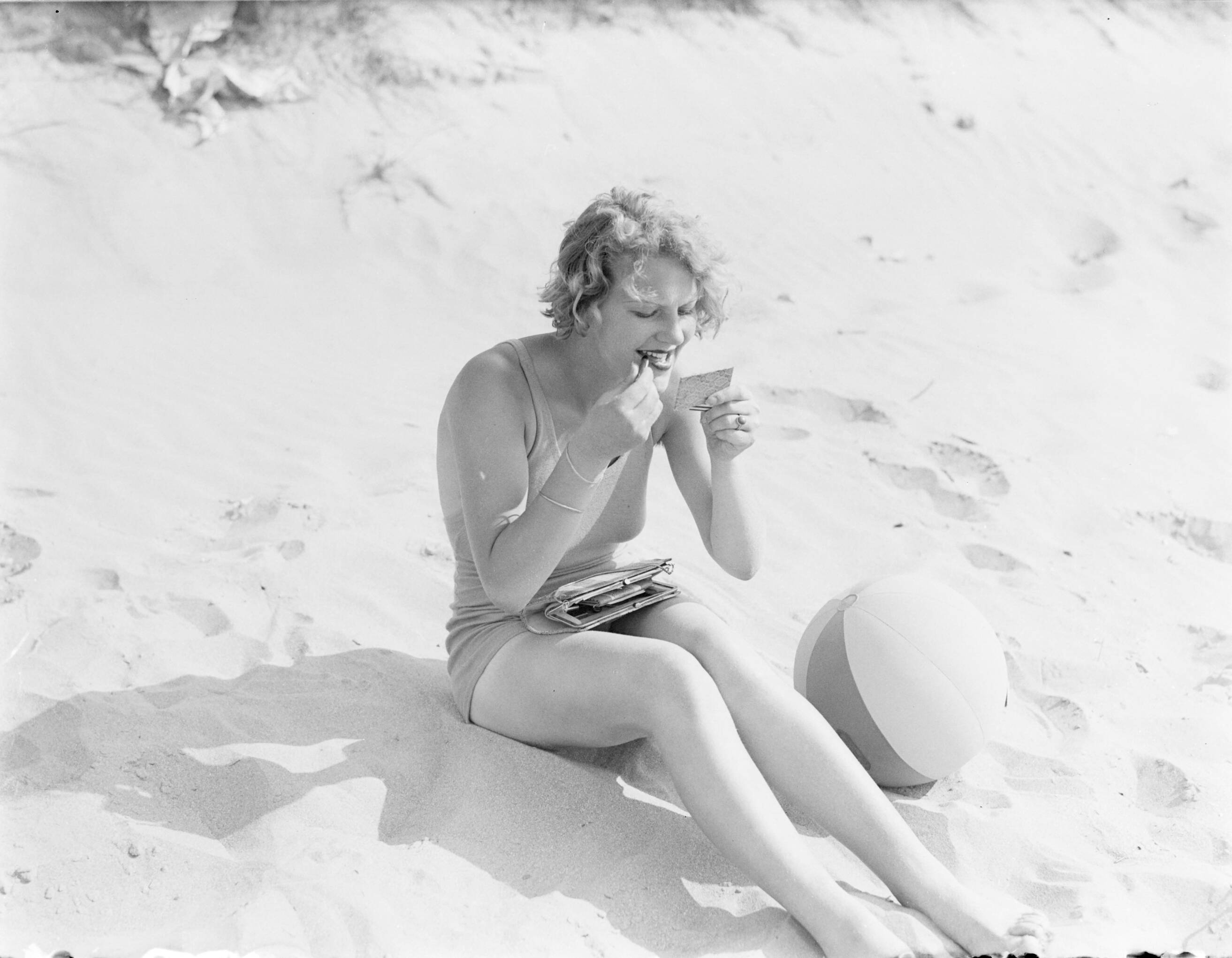 Reportage fotomodel — Model Eva Waldschmidt zit in het zand en stift haar lippen — Foto: Poll, Willem van de. Licentie: Set B: Rechtenvrij / Publiek Domein. Bron: Nationaal Archief. http://hdl.handle.net/10648/aea7f8b2-d0b4-102d-bcf8-003048976d84
