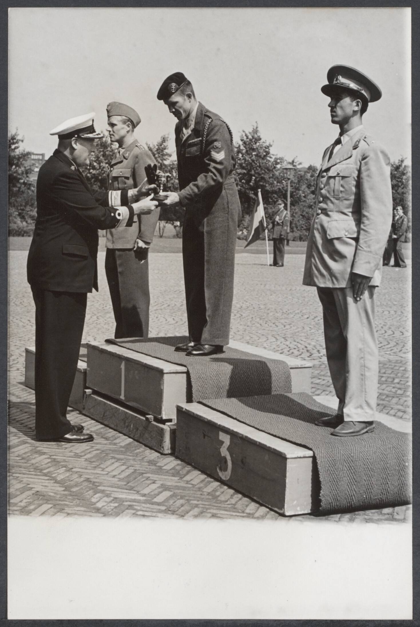 Ontspanning voor en door Nederlandse militairen - Nederland — Op de paradegrond van de Koninklijke Militaire Academie in Breda, werden zaterdag j.l. de prijzen uitgereikt aan de prijswinnaars in de Internationale Militaire Vijfkamp 1955, welke vanaf dinsdag in Breda is gehouden. De bevelhebber van de Nederlandse Zeestrijdkrachten, vice-admiraal A. de Booy, die alle prijzen uitreikte, overhandit hier de trofee voor het granaatwerpen aan de Nederlandse sergeant J. de Wijn. Op podium nr. 2 staat een Zweed, de luitenant Moberg en op no. 3 de Turkse luitenant Yilmaz — Foto: ANP / RVD of rechtenvrij. Licentie: Set B: Rechtenvrij / Publiek Domein. Bron: Nationaal Archief. http://hdl.handle.net/10648/388b1680-d7d5-71b0-d02f-ea7cbdccddd7