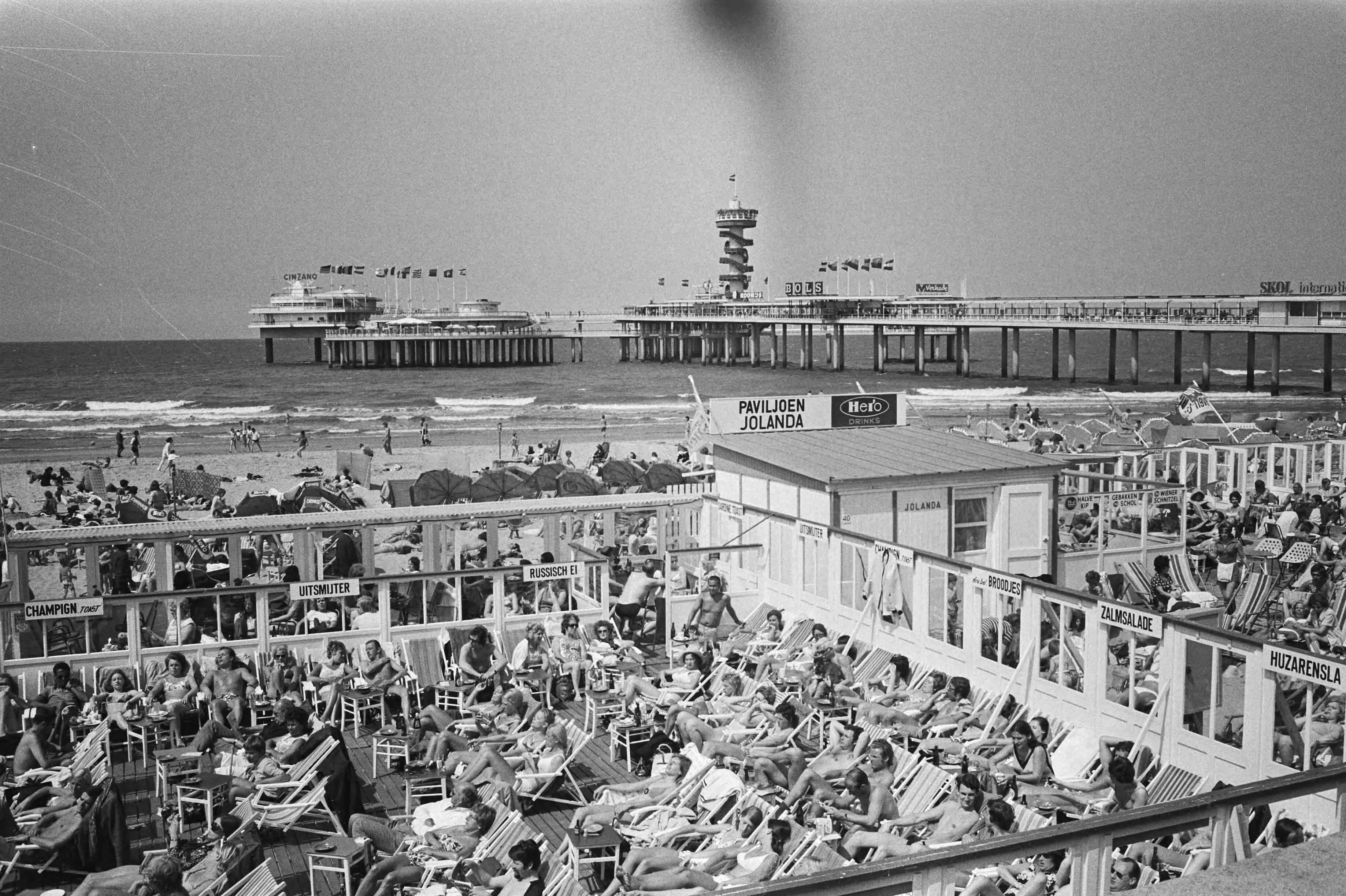 Drukte op terras strand Scheveningen — Volle terrassen met zonnende badgasten, op de achtergrond de pier — Foto: Fotograaf Onbekend / Anefo. Licentie: Set B: Rechtenvrij / Publiek Domein. Bron: Nationaal Archief. http://hdl.handle.net/10648/abc00388-d0b4-102d-bcf8-003048976d84