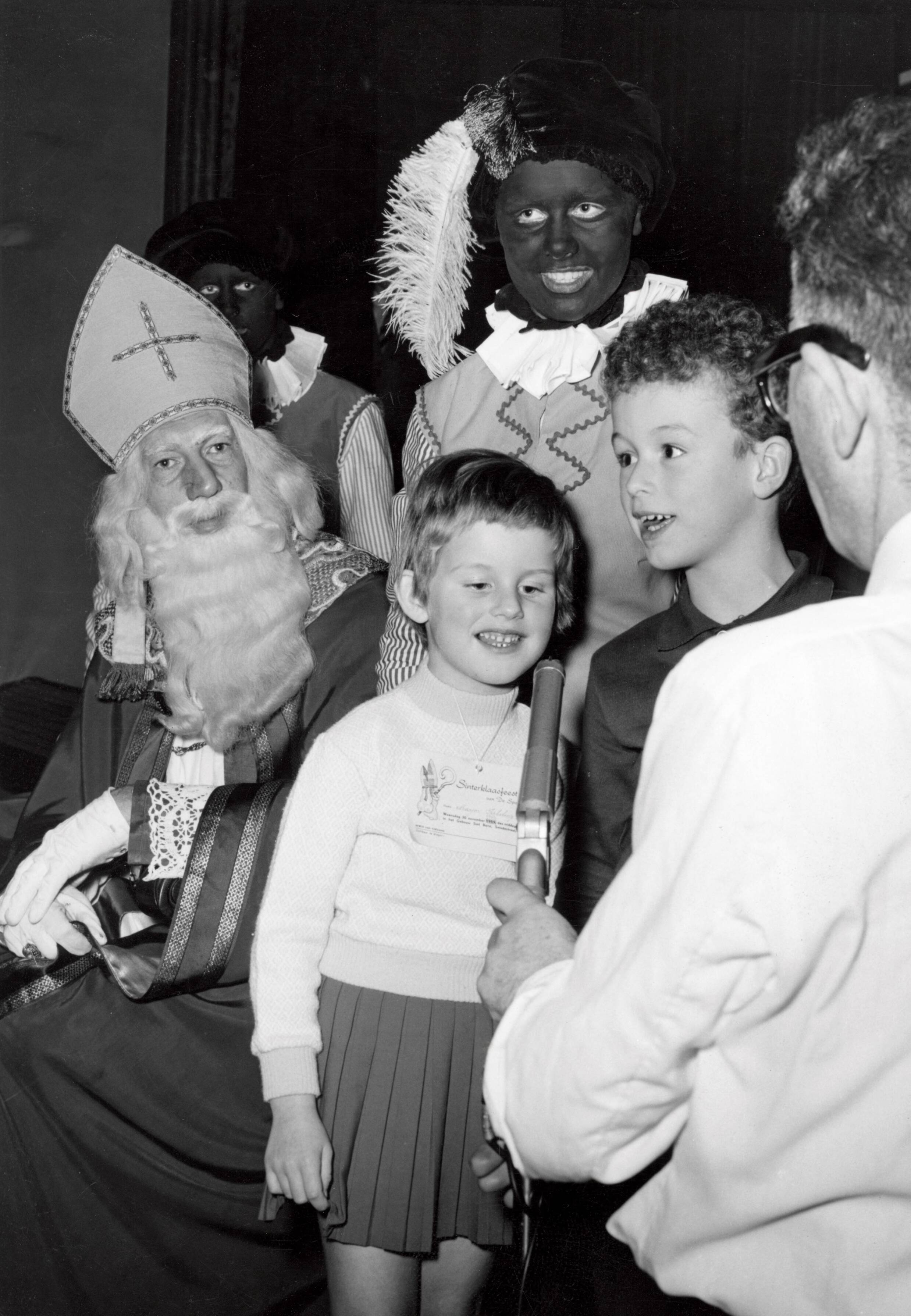 Collectie nader te bepalen — [Sinterklaasfeest N.V. Drukkerij De Spaarnestad in Haarlem] [twee kinderen spreken in een voor hen gehouden microfoon, met achter hun Sinterklaas en Zwarte Piet] — Foto: Fotograaf Onbekend / Fotobureau onbekend / Spaarnestad Photo (05887). Licentie: Set B: Rechtenvrij / Publiek Domein. Bron: Nationaal Archief. http://hdl.handle.net/10648/35429da6-4050-a382-e063-6df0900a6686