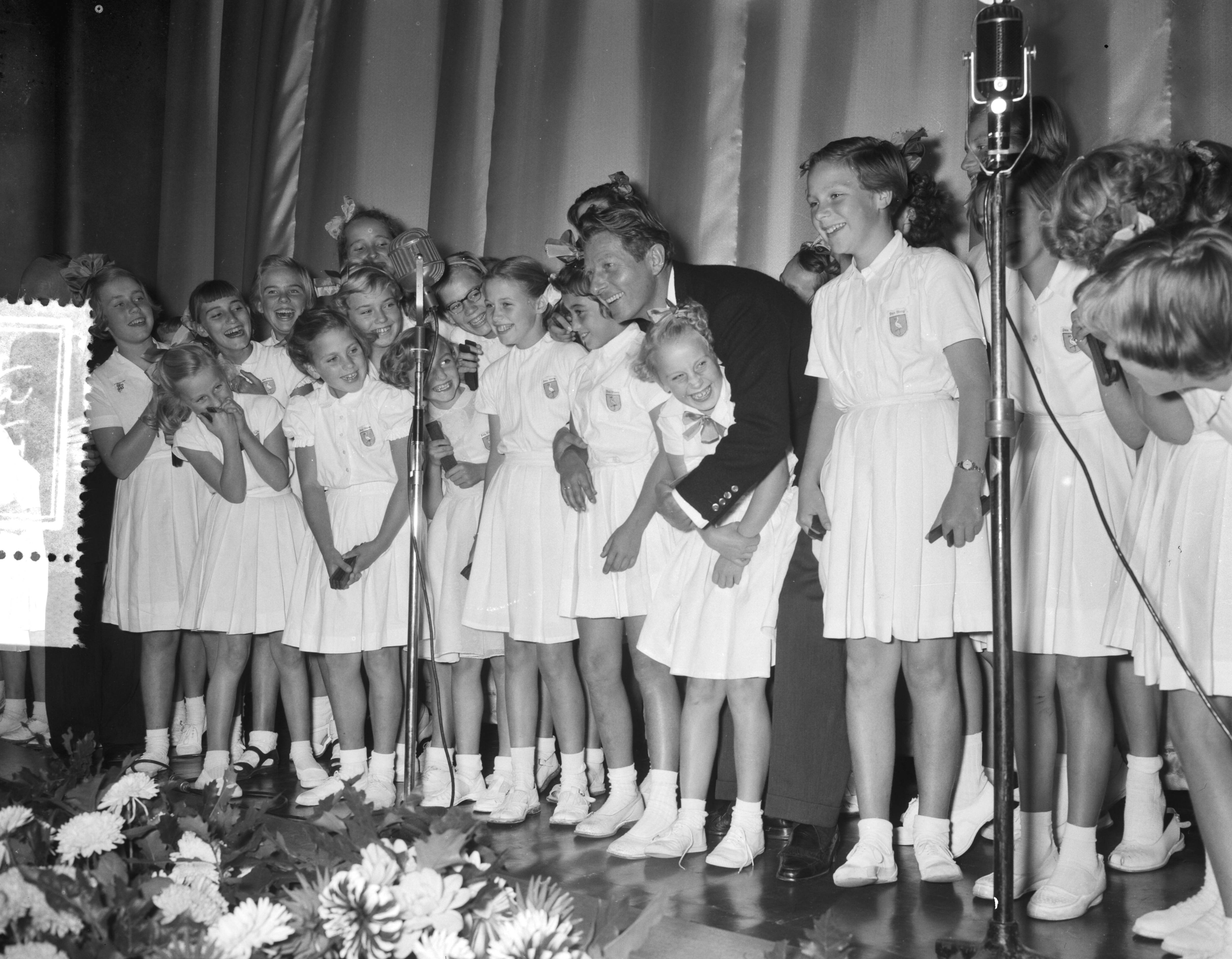 Fotocollectie Anefo — Première van de UNICEF-film "Kinderogen lachen weer" in het Metropole-Tuschinski theater te Den Haag. Danny Kaye op het toneel met het kinderkoor "De Haagse Ooievaartjes" — Foto: Noske, J.D. / Anefo. Licentie: Set B: Rechtenvrij / Publiek Domein. Bron: Nationaal Archief. http://hdl.handle.net/10648/a93c579c-d0b4-102d-bcf8-003048976d84