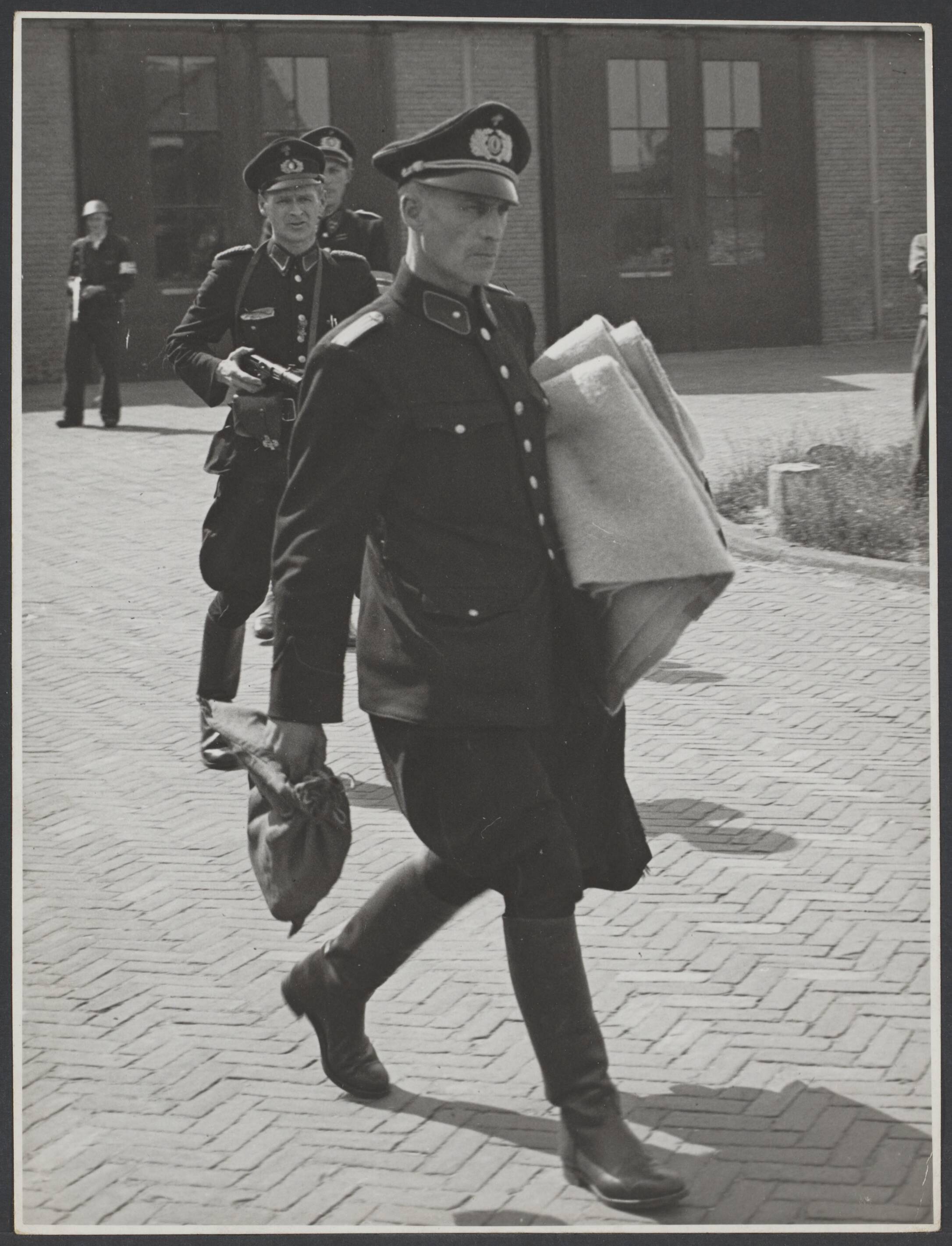 Collaboratie Nederland. — Haarlem [man in uniform [staatspolitie?] loopt - onder schot gehouden door collega's?- met dekens over een binnenplaats. In de achtergrond een bewapende B.S.-er] — Foto: Nationaal Foto Persbureau (Stevens & Magielsen) / Rechtenvrij. Licentie: Set B: Rechtenvrij / Publiek Domein. Bron: Nationaal Archief. http://hdl.handle.net/10648/0cde9240-7541-2a28-7c19-8be05e17acd4