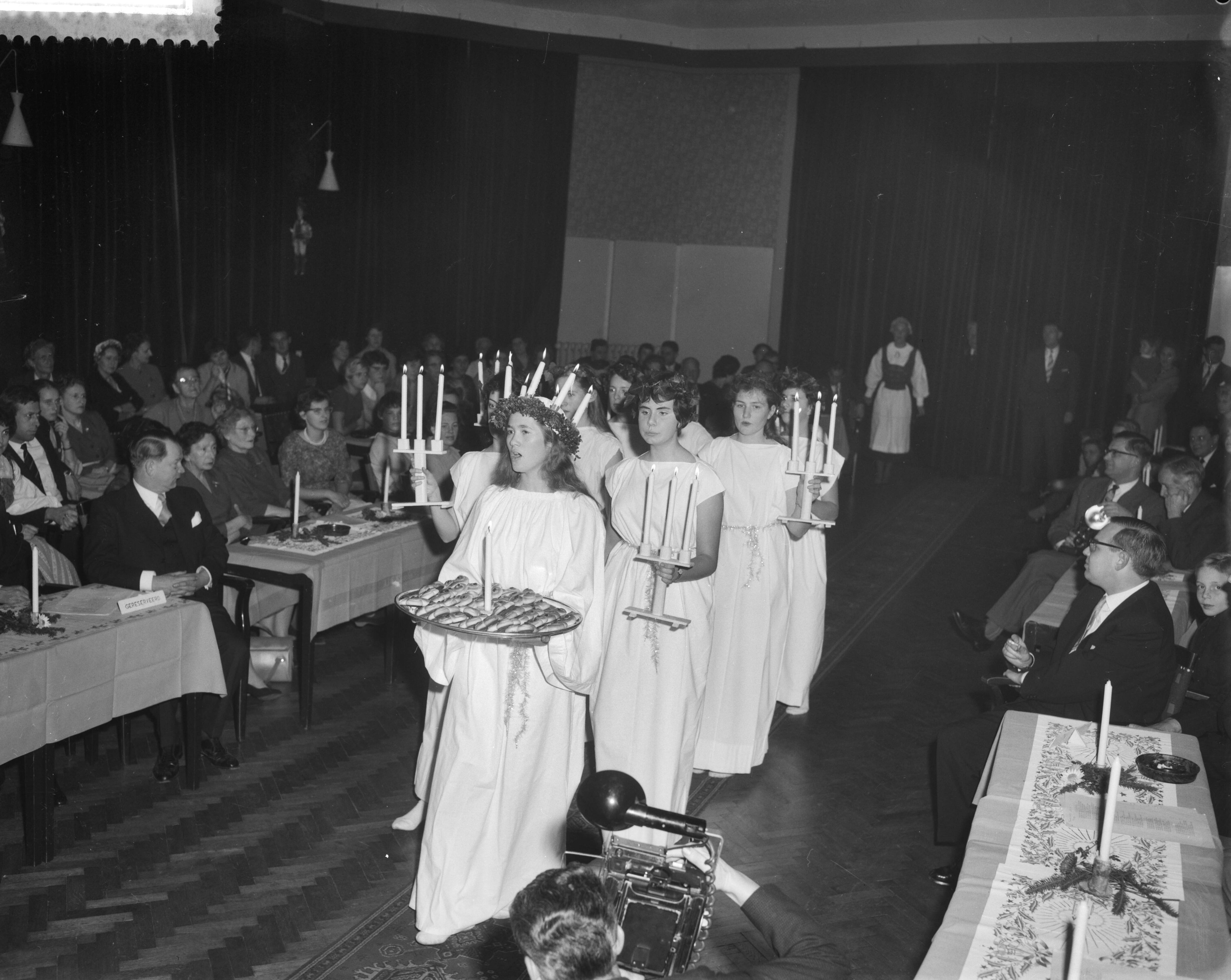 Fotocollectie Anefo — Viering van het Luciafeest door de Zweeds Nederlandse Vereniging in de Dierentuin te Den Haag, Lucia (de 18-jarige Helbertijn Küller uit Den Haag) gevolgd door "ternen" komt de zaal binnen — Foto: Behrens, Herbert F. / Anefo. Licentie: Set B: Rechtenvrij / Publiek Domein. Bron: Nationaal Archief. http://hdl.handle.net/10648/a9a3aed8-d0b4-102d-bcf8-003048976d84