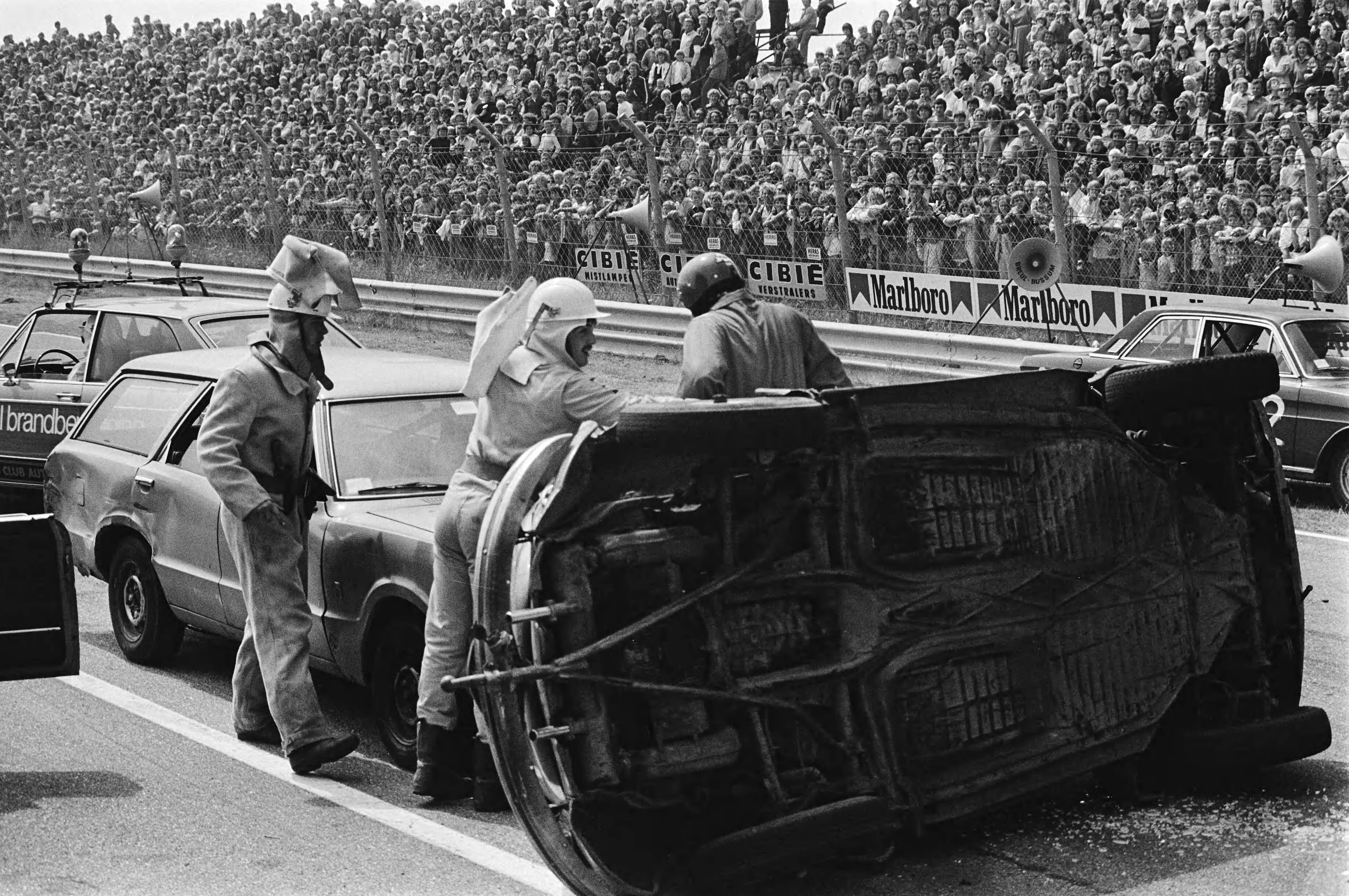 "Ter land, ter zee en in de lucht", opnamen voor het TROS TV programma wedstrijd achteruitrijden op het circuit van Zandvoort — Mannen bij een gekantelde auto — Foto: Suyk, Koen / Anefo. Licentie: Set B: Rechtenvrij / Publiek Domein. Bron: Nationaal Archief. http://hdl.handle.net/10648/acce2444-d0b4-102d-bcf8-003048976d84