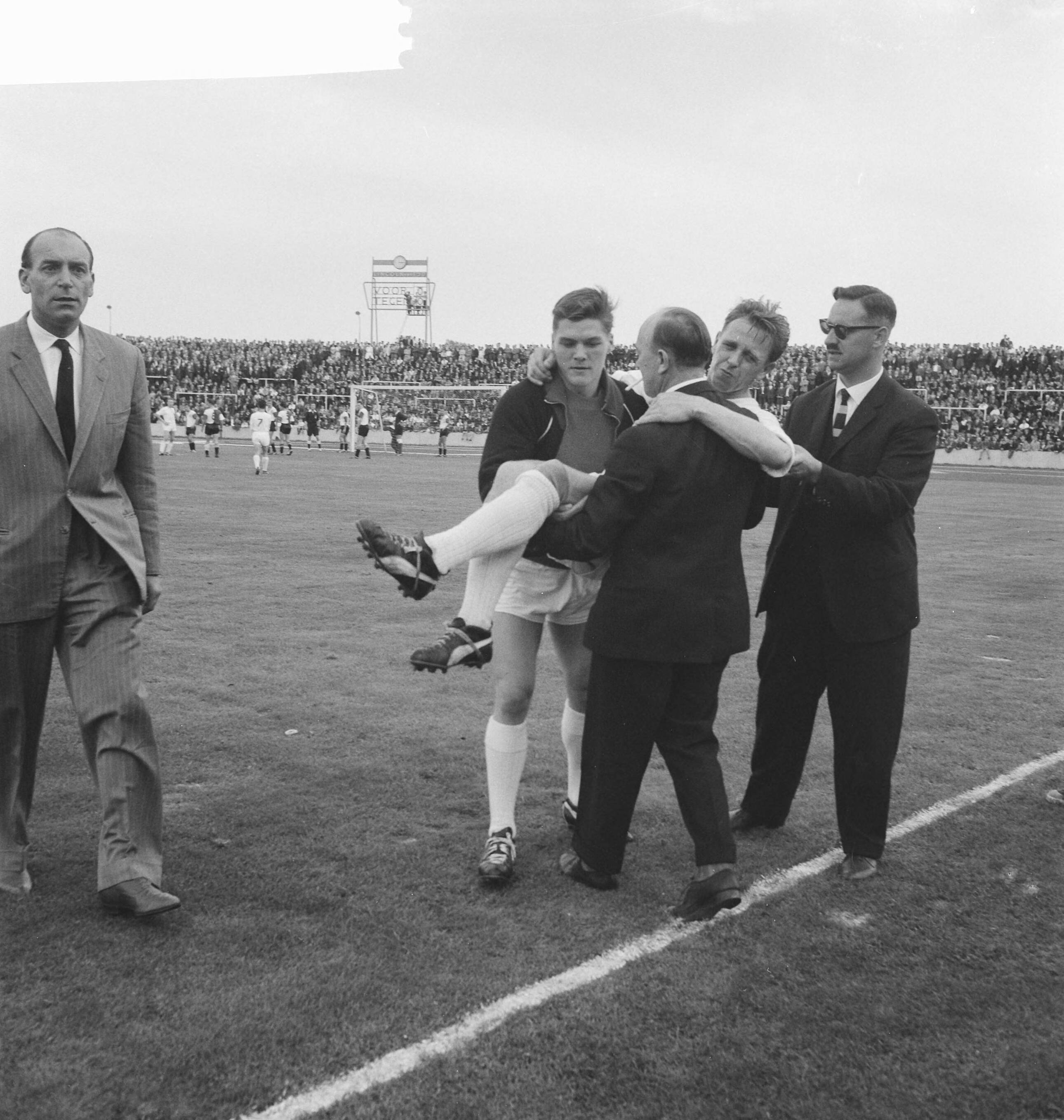 Fotocollectie Anefo — Voetbal. MVV tegen Feijenoord 0-0. Frits Stevens van MVV wordt geblesseerd van het veld gedragen, o.a. door medespeler Chris Coenen — Foto: Koch, Eric / Anefo. Licentie: Set B: Rechtenvrij / Publiek Domein. Bron: Nationaal Archief. http://hdl.handle.net/10648/a9e49c2c-d0b4-102d-bcf8-003048976d84