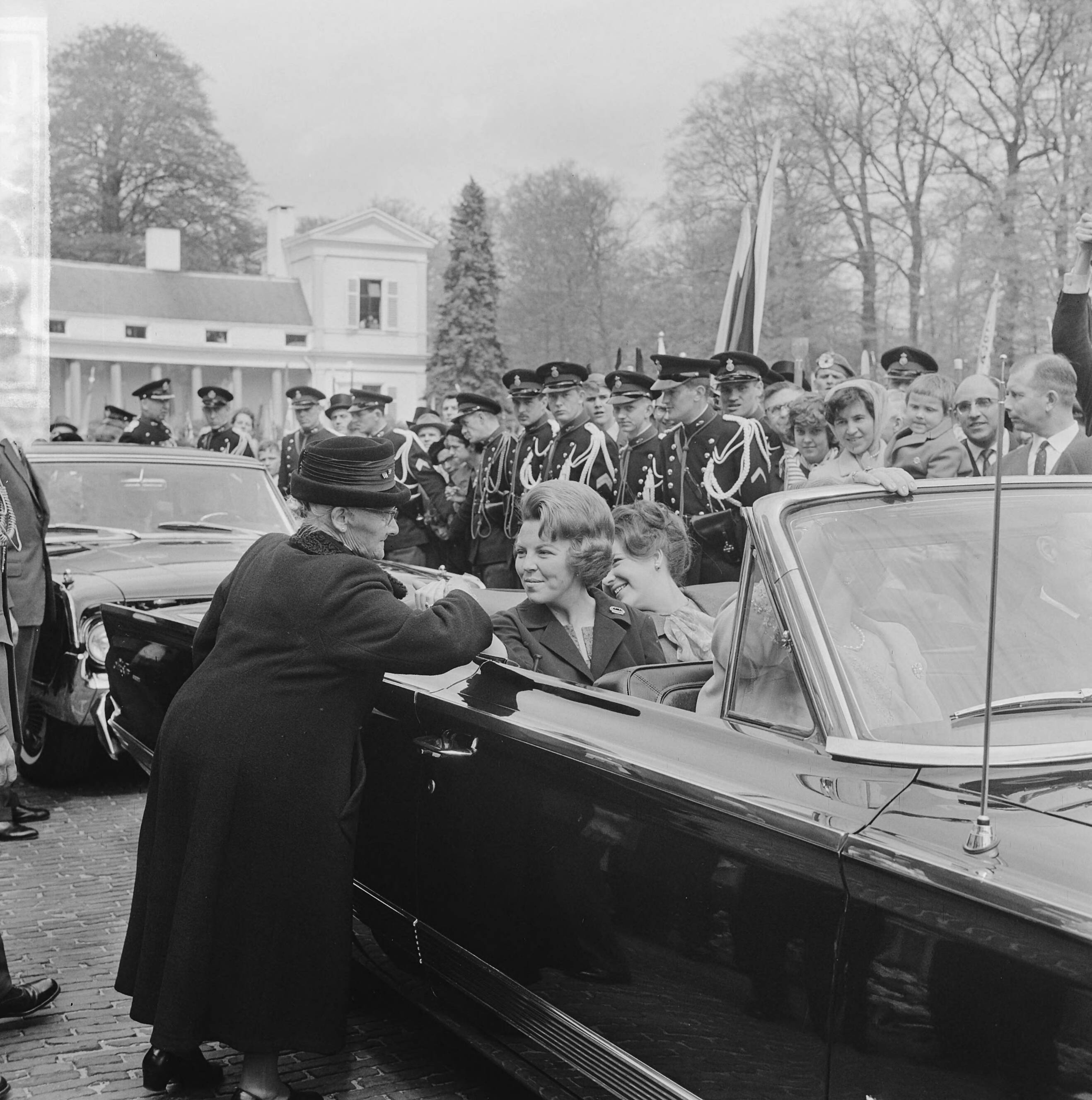 Koninginnedag 1964 — Defilé Soestdijk, tijdens autorit door paleistuin, een bejaarde vrouw houdt de pols van prinses Beatrix vast — Foto: Koch, Eric / Anefo. Licentie: Set B: Rechtenvrij / Publiek Domein. Bron: Nationaal Archief. http://hdl.handle.net/10648/aa66c742-d0b4-102d-bcf8-003048976d84