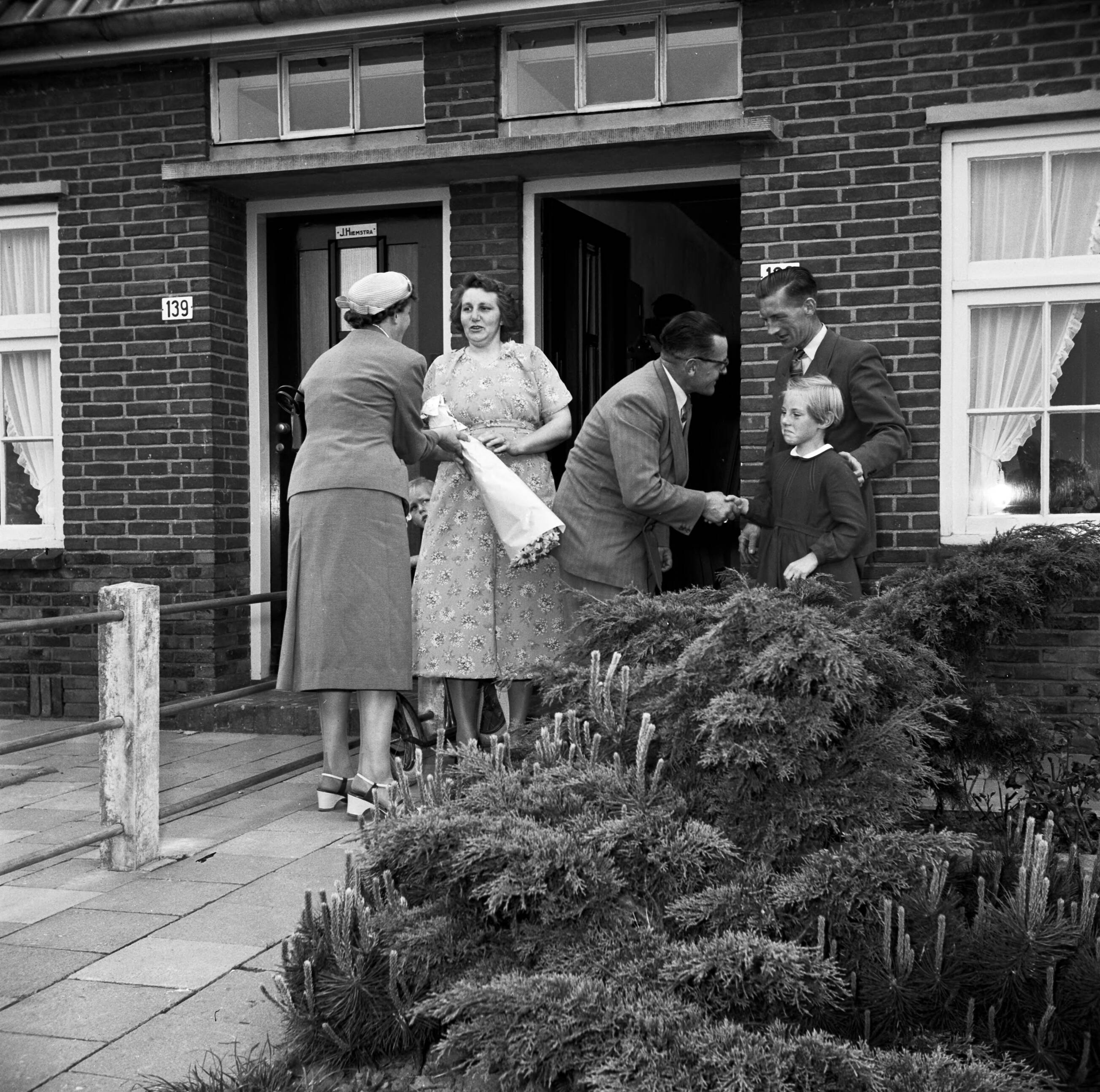 Fotocollectie Van de Poll — Gerard de Jong en zijn echtgenote bij de voordeur van hun woning. Ynse Postma schudt de hand van hun dochtertje Marietje. Links De Jongs echtgenote die een boeket in de hand houdt en met Postma's echtgenote spreekt — Foto: Poll, Willem van de. Licentie: Set B: Rechtenvrij / Publiek Domein. Bron: Nationaal Archief. http://hdl.handle.net/10648/aecce226-d0b4-102d-bcf8-003048976d84