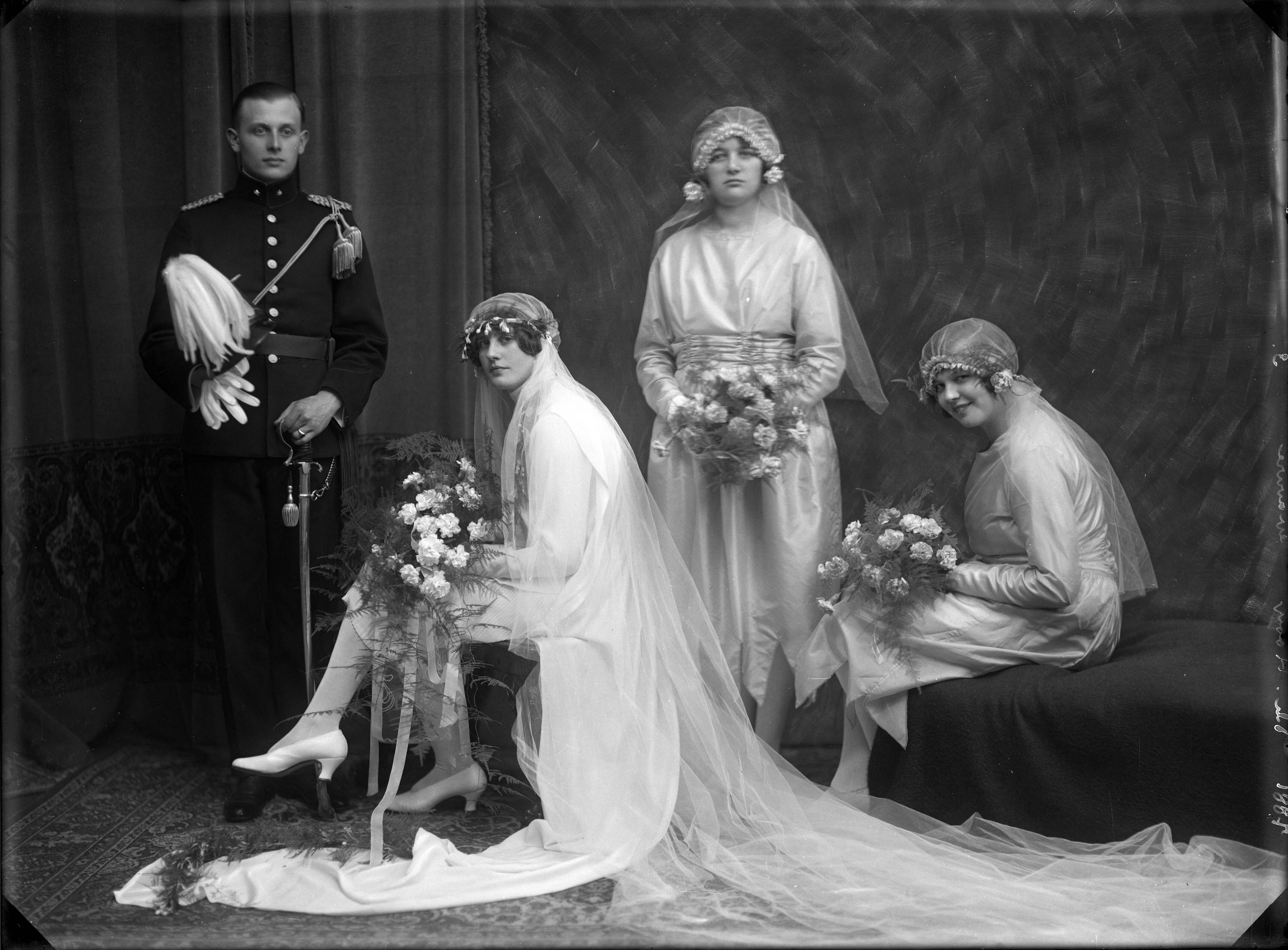Berg, van den (BERG 050) — Studio-opnames. Een bruidspaar met twee volwassen bruidsmeisjes. De bruidegom in officiers-uniform, de bruid zittend in een korte witte jurk met lange sleep van satijn, een hoofdtooi van tule met daaraan een lange sleep. De bruidsmeisjes in een gladde witte jurk met eveneens een hoofdtooi van tule . Met bruidsboeketten van witte rozen. 7881 Th. Hooy gravure 3.Haarlem, Nederland. 1918-1930 — Foto: Berg, Th.C.J. van den / Spaarnestad Photo (05887). Licentie: Set B: Rechtenvrij / Publiek Domein. Bron: Nationaal Archief. http://hdl.handle.net/10648/35429da7-c5b7-a382-e063-6df0900a6686