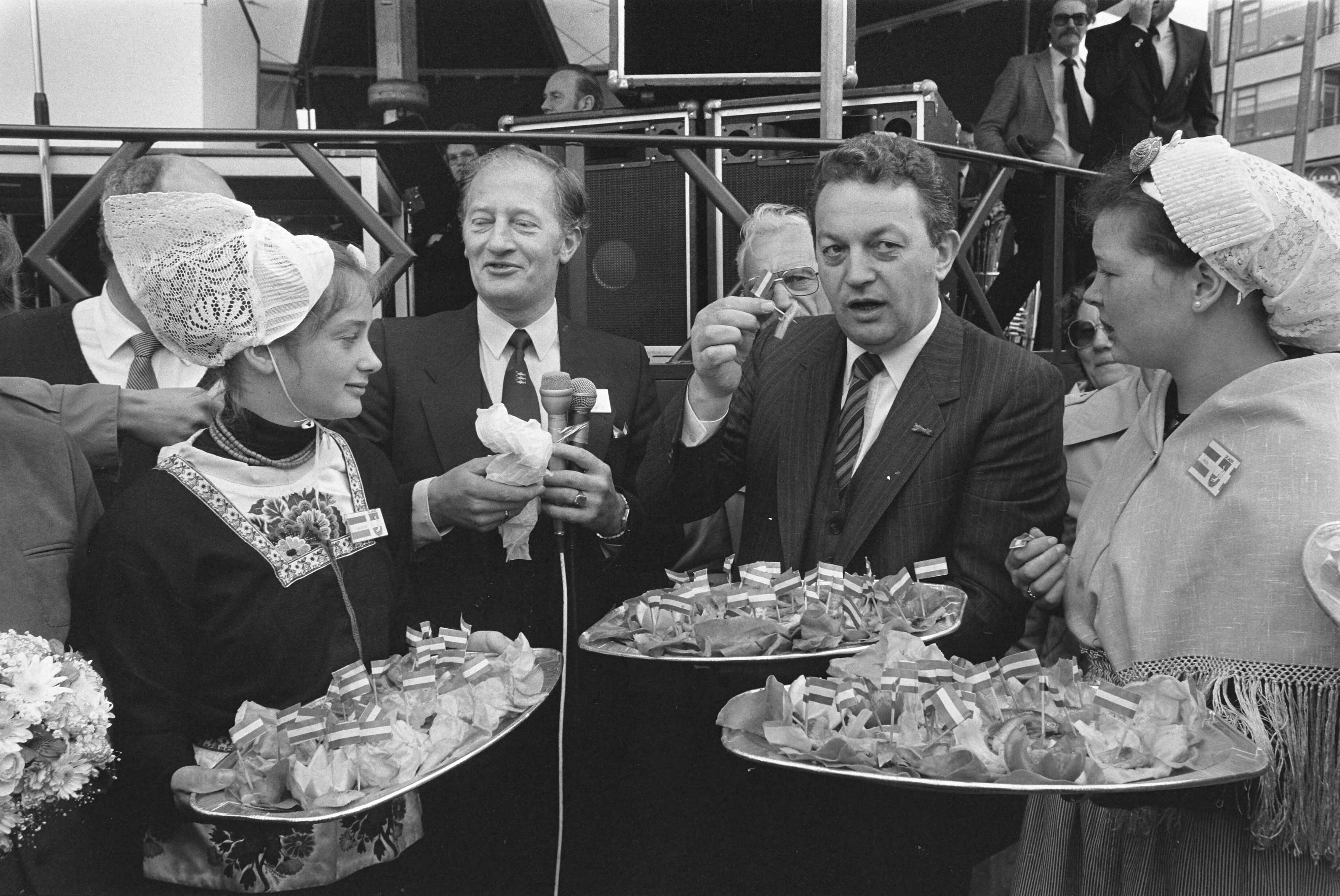 Vlaggetjesdag in Scheveningen 1983 — Minister Braks (Landbouw en Visserij) krijgt haring aangeboden door een vrouw in klederdracht — Foto: Croes, Rob C. / Anefo. Licentie: Set B: Rechtenvrij / Publiek Domein. Bron: Nationaal Archief. http://hdl.handle.net/10648/ad29db2c-d0b4-102d-bcf8-003048976d84