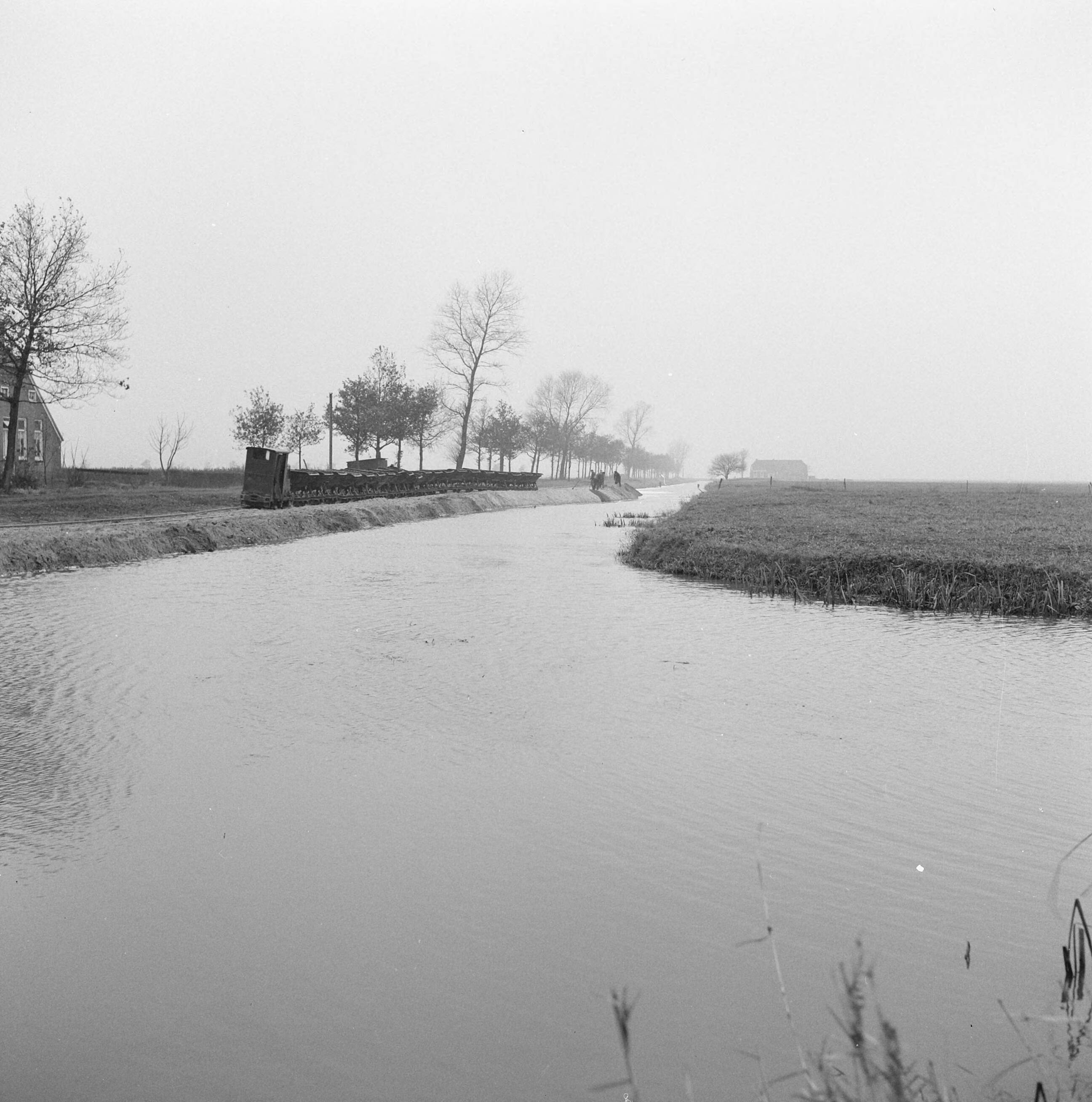 Fotocollectie Nederlandse Heidemaatschappij — Het dempen van het hoofdkanaal bijlangs de benedenste Veenweg. De bestaande sintelweg (links op de foto vlak voor de boerderijen langs) en het kanaal vervallen en wordt gespit tot bouwland. De nieuwe wegen komen grotendeels over de te dempen kanalen te lopen. Het zandkunst is hier te zien rechts van de houten schuilkeet. De brug recht overbrugt de zijwijkvan de fam. Maarsingh en Meursing. Deze wijk wordt afgesloten door de aanleg van de weg en daarna gedempt — Foto: Wüllschleger, [...] / Heidemij. Licentie: Set B: Rechtenvrij / Publiek Domein. Bron: Nationaal Archief. http://hdl.handle.net/10648/ae71c972-d0b4-102d-bcf8-003048976d84