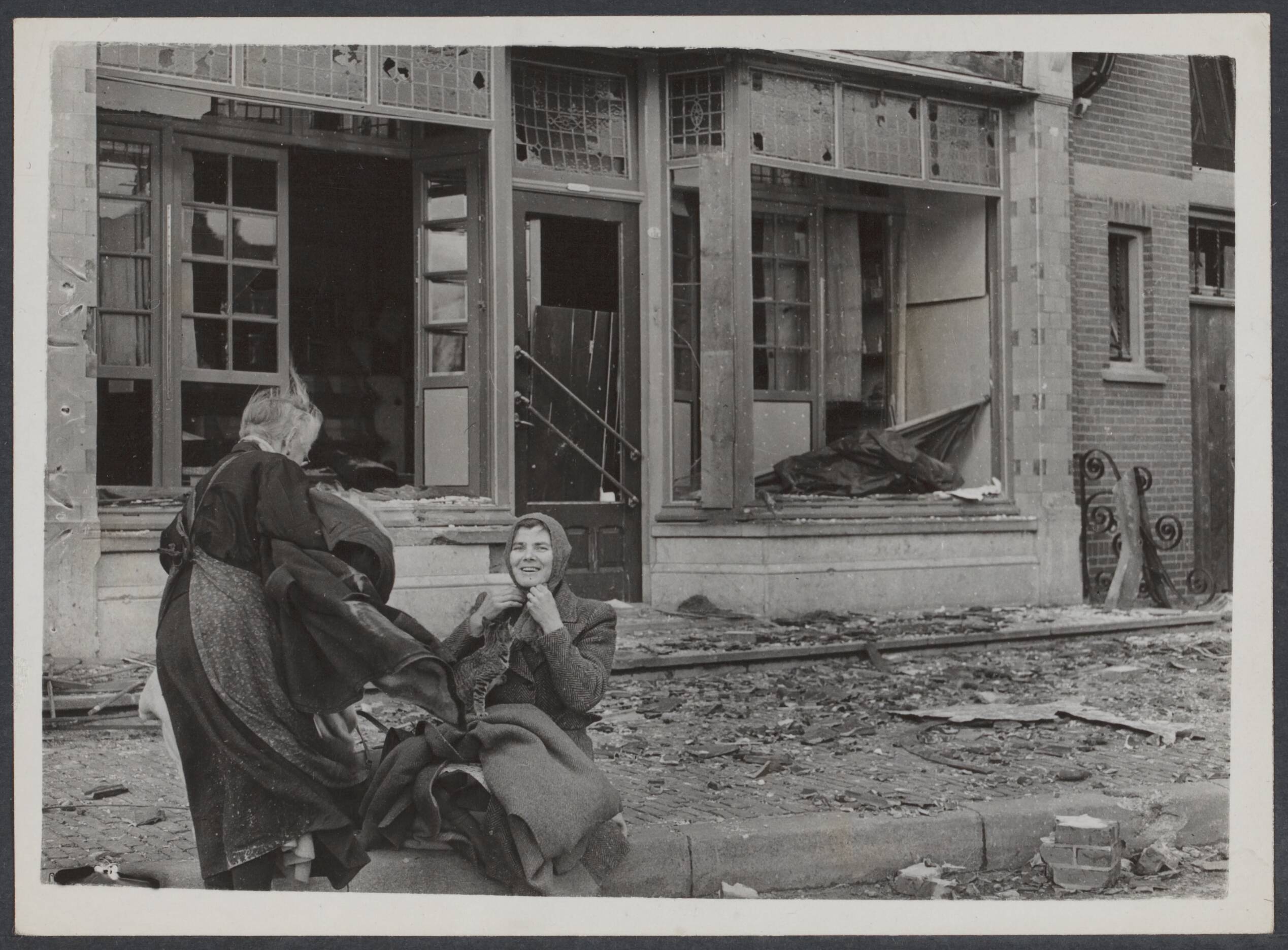 Nederland front, 1944 — In het Brabantse dorpje Wagenberg zitten twee vrouwen en een kat buiten voor hun verwoeste winkel — Foto: Keystone / Rechtenvrij. Licentie: Set B: Rechtenvrij / Publiek Domein. Bron: Nationaal Archief. http://hdl.handle.net/10648/d15580af-a104-bfaf-70c3-197a04c2ebad