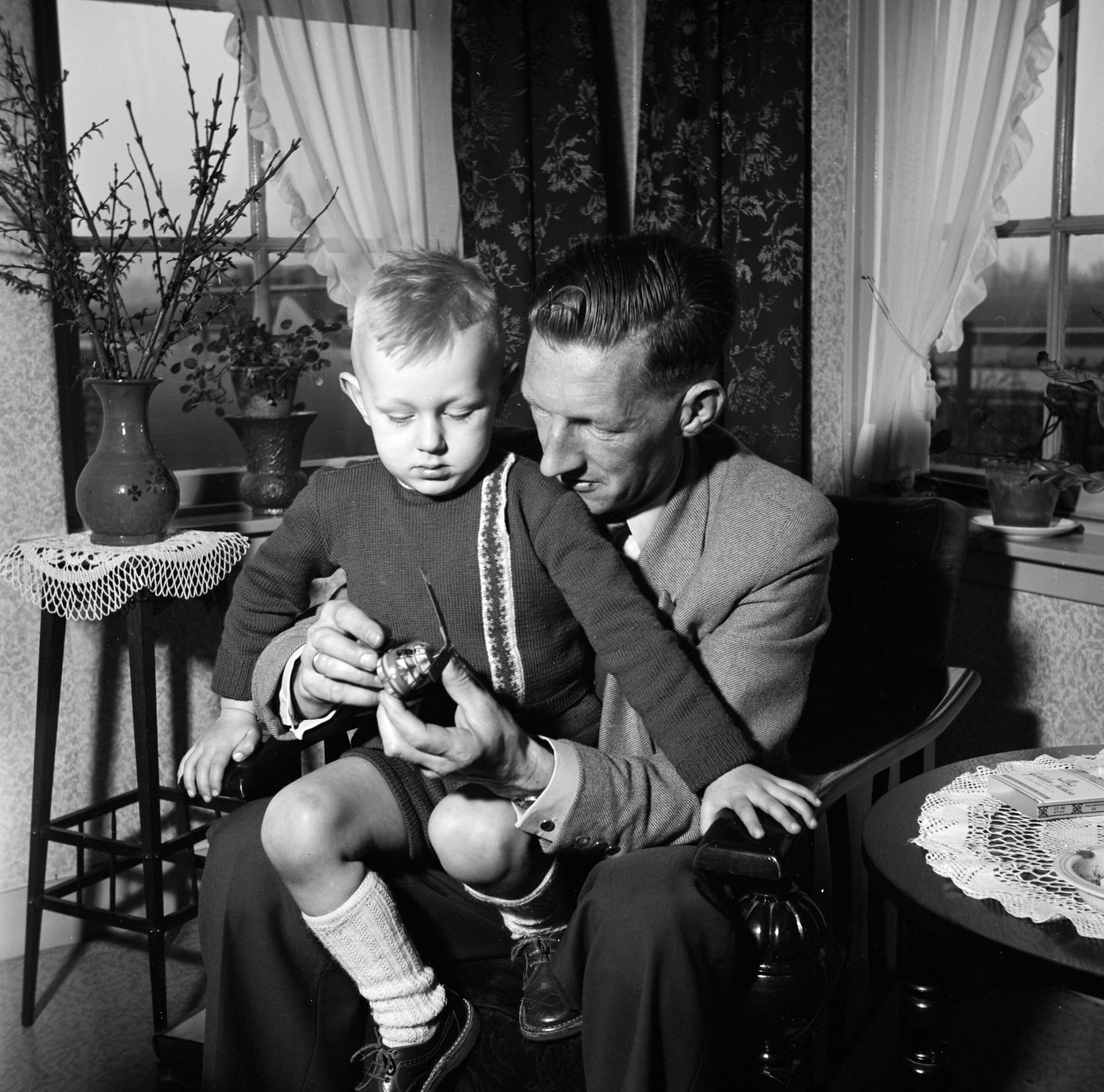 Fotocollectie Van de Poll — Gerard met zijn vierjarig zoontje Gerrie op schoot met een speelgoed helikopter. Links en bloementafeltje met een vaas met uitbottende takken — Foto: Poll, Willem van de. Licentie: Set B: Rechtenvrij / Publiek Domein. Bron: Nationaal Archief. http://hdl.handle.net/10648/aeccd0b0-d0b4-102d-bcf8-003048976d84