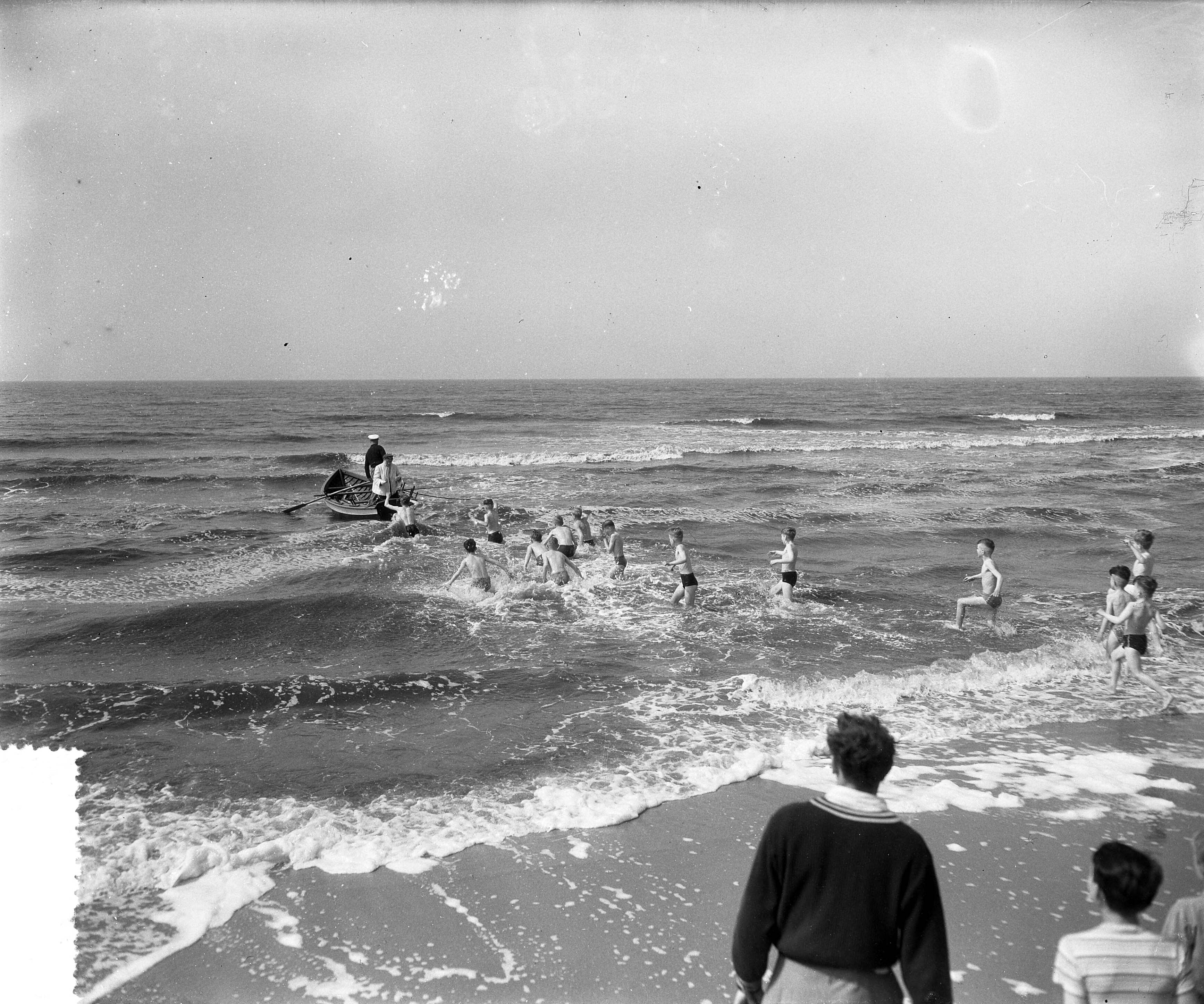 Fotocollectie Anefo — "Wedloop naar zee" te Scheveningen [een groep jongens in zwembroek rennen de zee in, naar een roeiboot in de branding, waar een vrouw klaarstaat met een voorwerp] — Foto: Rossem, Wim van / Anefo. Licentie: Set B: Rechtenvrij / Publiek Domein. Bron: Nationaal Archief. http://hdl.handle.net/10648/a935f76c-d0b4-102d-bcf8-003048976d84