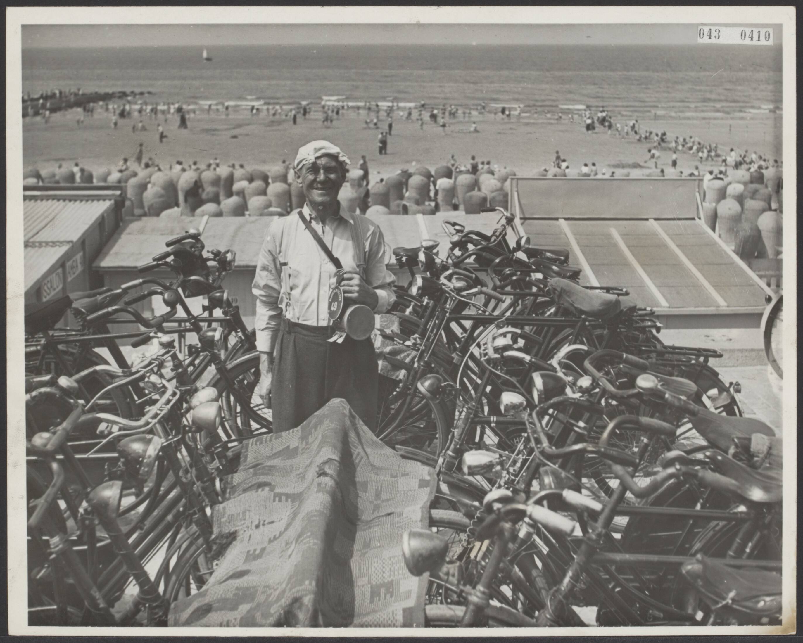 Fotocollectie Elsevier Binnenland — [Serie "Summum". over vermaak in Scheveningen] 2. Glunderend lacht u de man van de fietsenstalling tegen — Foto: Associated Correspondents. Licentie: Set B: Rechtenvrij / Publiek Domein. Bron: Nationaal Archief. http://hdl.handle.net/10648/adf8e868-d0b4-102d-bcf8-003048976d84