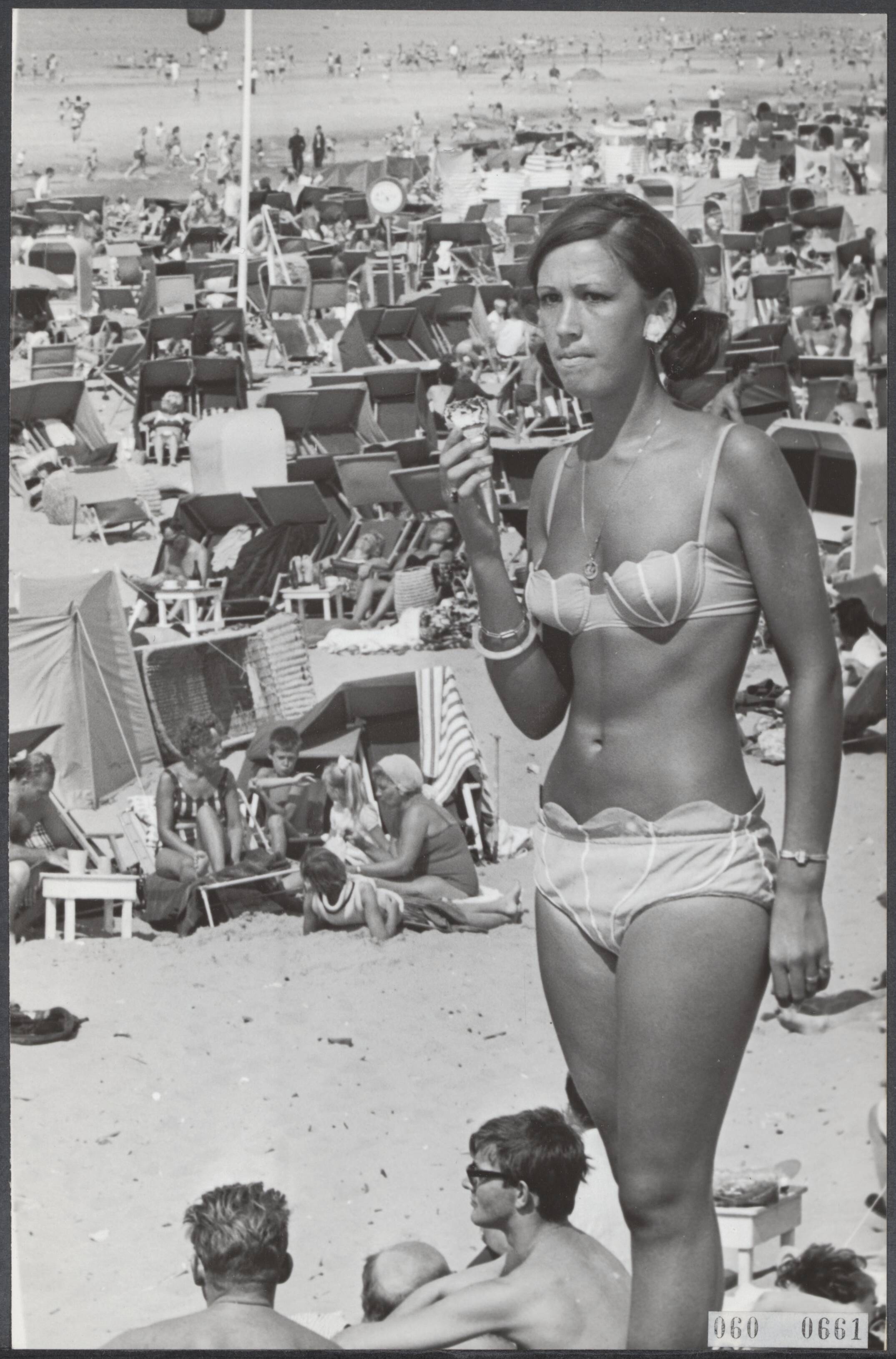 Fotocollectie Elsevier Binnenland — Stralend weer op Zandvoort. Vrouw in bikini met een ijsje. Achter haar een vol strand — Foto: Kroon, Ron / Anefo. Licentie: Set B: Rechtenvrij / Publiek Domein. Bron: Nationaal Archief. http://hdl.handle.net/10648/ae0df55a-d0b4-102d-bcf8-003048976d84