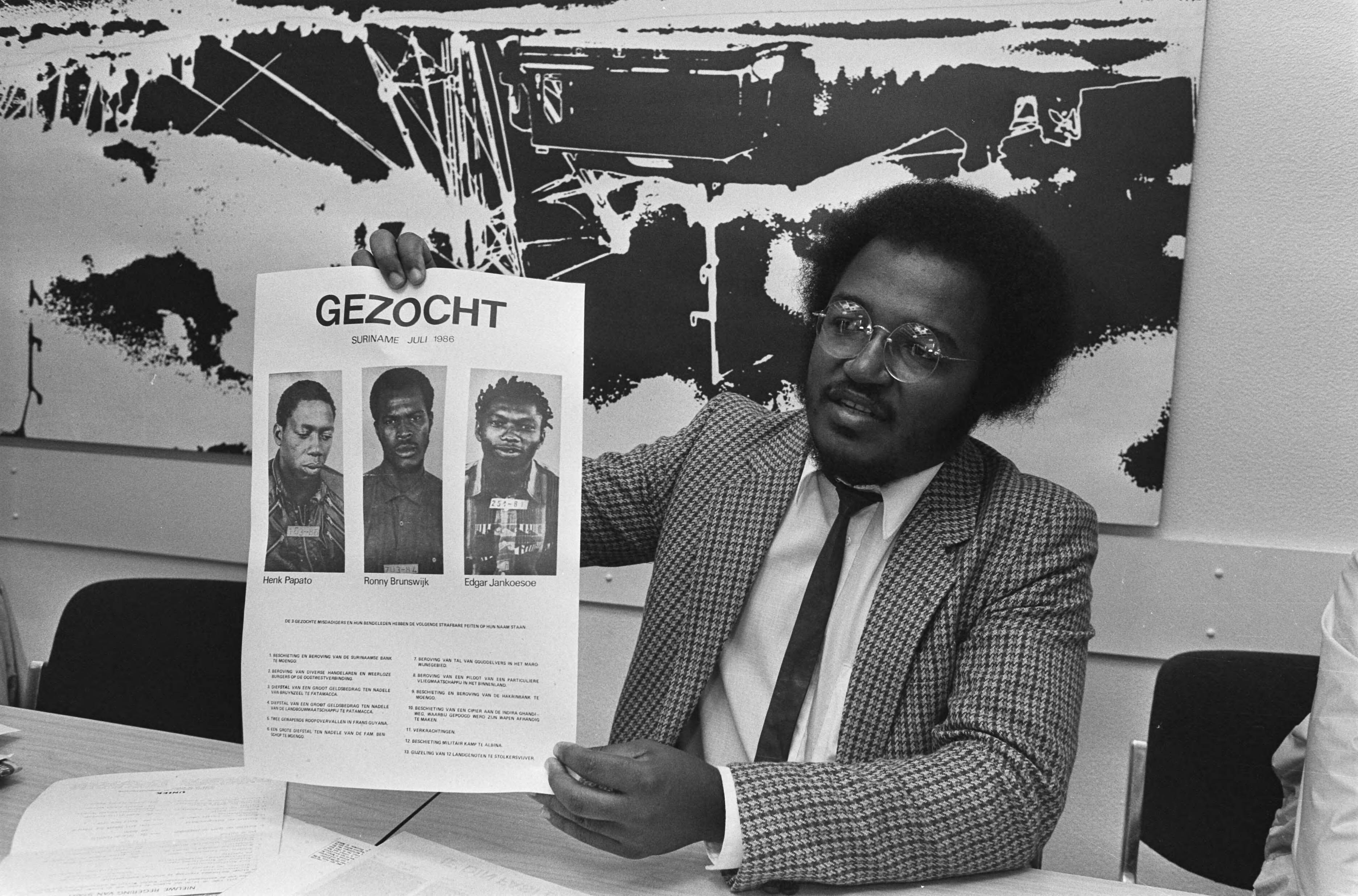 Fotocollectie Anefo — Pro Bouterse bijeenkomst in Den Haag ; Ludwig van Mulier (Liga Gen. Patriotten) met affiche met gezochte rebellenleiders — Foto: Croes, Rob C. / Anefo. Licentie: Set B: Rechtenvrij / Publiek Domein. Bron: Nationaal Archief. http://hdl.handle.net/10648/ad55d632-d0b4-102d-bcf8-003048976d84