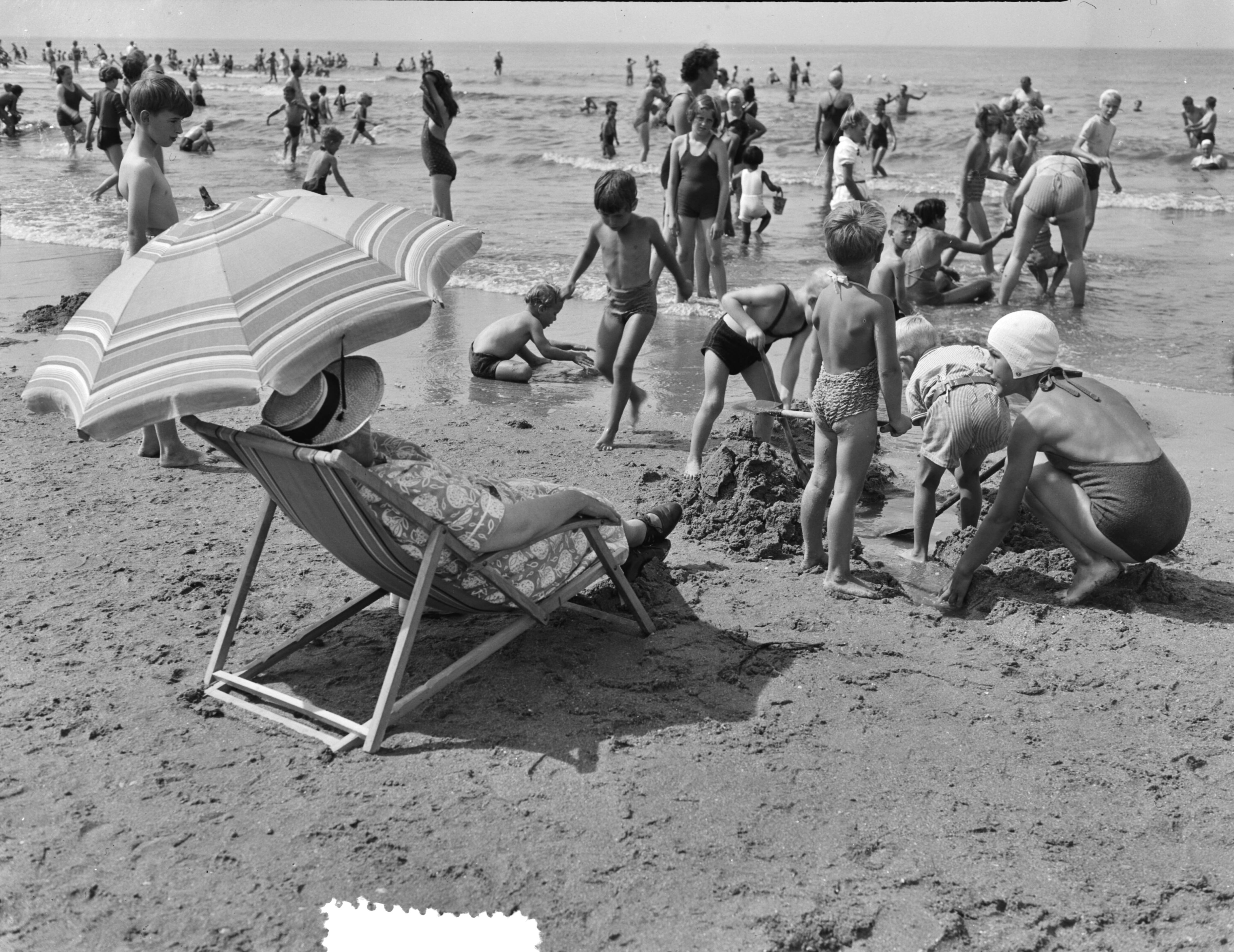 Fotocollectie Anefo — Hete dag aan het strand van Scheveningen. Drukte aan de vloedlijn [vrouw in strandstoel met parasol] — Foto: Duinen, G.W. van / Anefo. Licentie: Set B: Rechtenvrij / Publiek Domein. Bron: Nationaal Archief. http://hdl.handle.net/10648/a910eab2-d0b4-102d-bcf8-003048976d84
