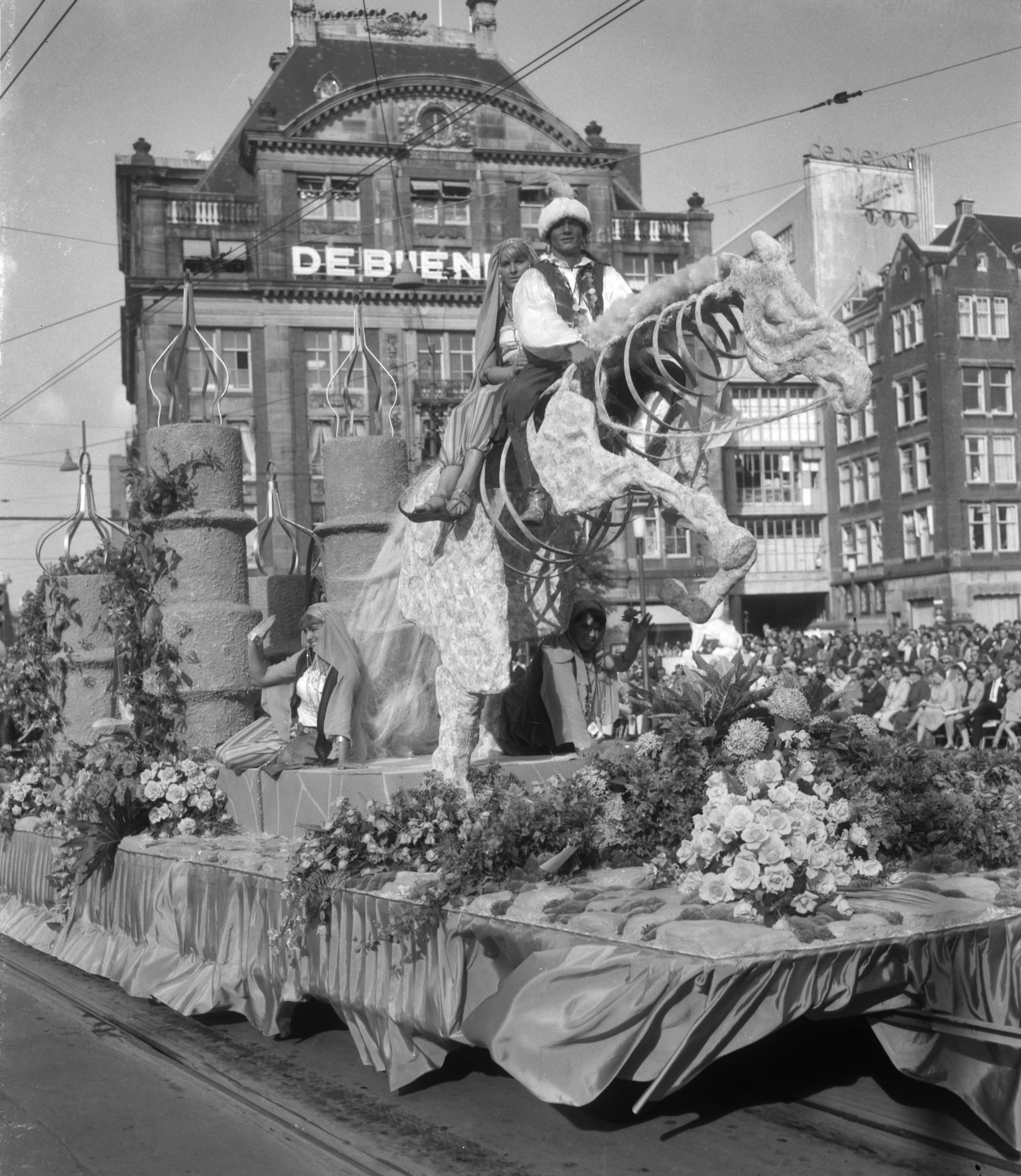 Bloemencorso van Aalsmeer te Amsterdam — Een praalwagen bij de Dam te Amsterdam met op de achtergrond het warenhuis de Bijenkorf — Foto: Bilsen, Joop van / Anefo. Licentie: Set B: Rechtenvrij / Publiek Domein. Bron: Nationaal Archief. http://hdl.handle.net/10648/aafae9d6-d0b4-102d-bcf8-003048976d84