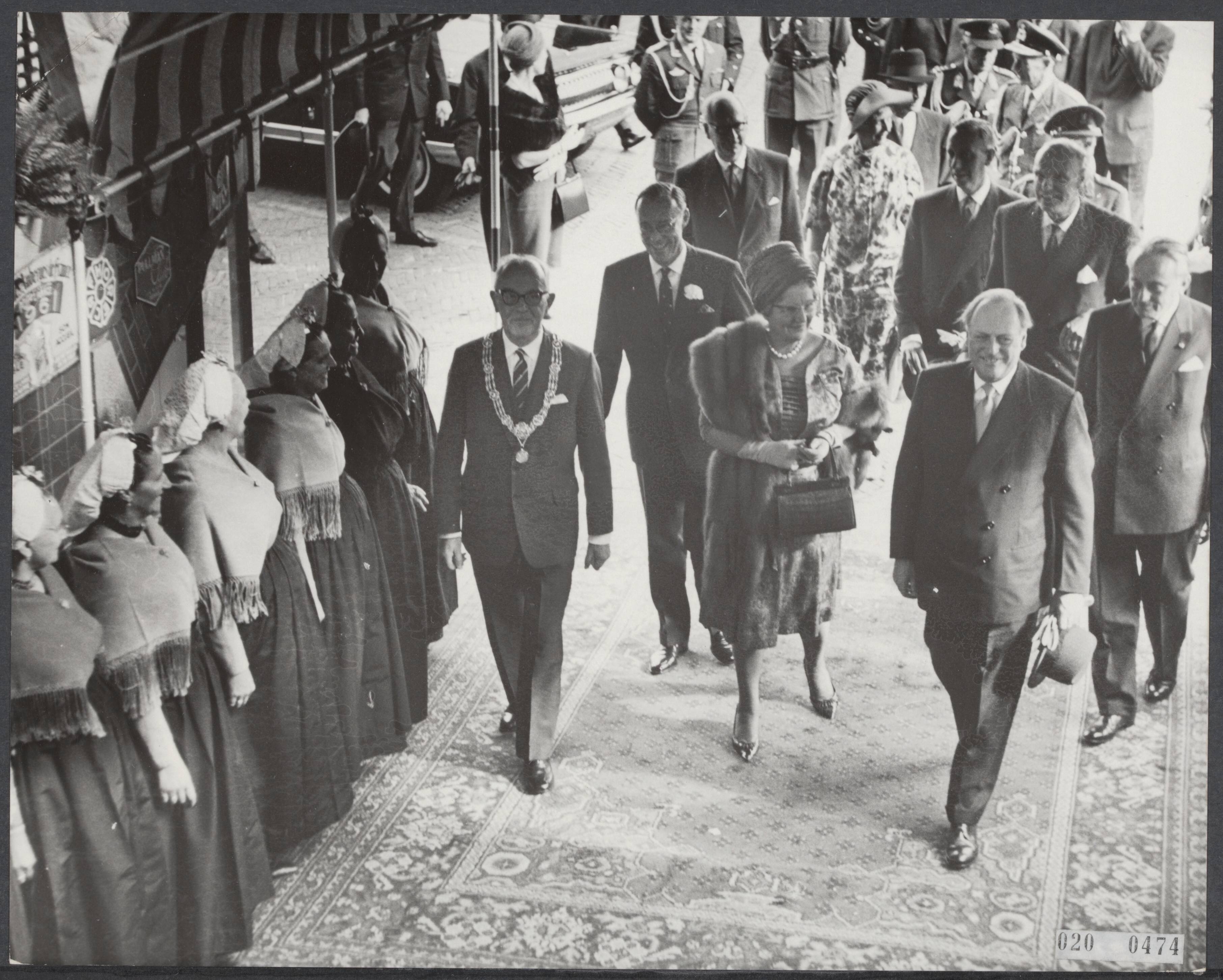 Fotocollectie Elsevier Binnenland — [Staatsbezoek van koning Olav van Noorwegen aan Nederland] Koning Olav van Noorwegen, koningin Juliana en prins Bernhard wonen in het Kurhaus een speciale voorstelling bij van Bert Haanstra´s film alleman. De koninklijke gasten betreden het Kurhaus door langs twee rijen Scheveningse vissersvrouwen door te lopen — Foto: Koch, Eric / Anefo. Licentie: Set B: Rechtenvrij / Publiek Domein. Bron: Nationaal Archief. http://hdl.handle.net/10648/adcaf250-d0b4-102d-bcf8-003048976d84