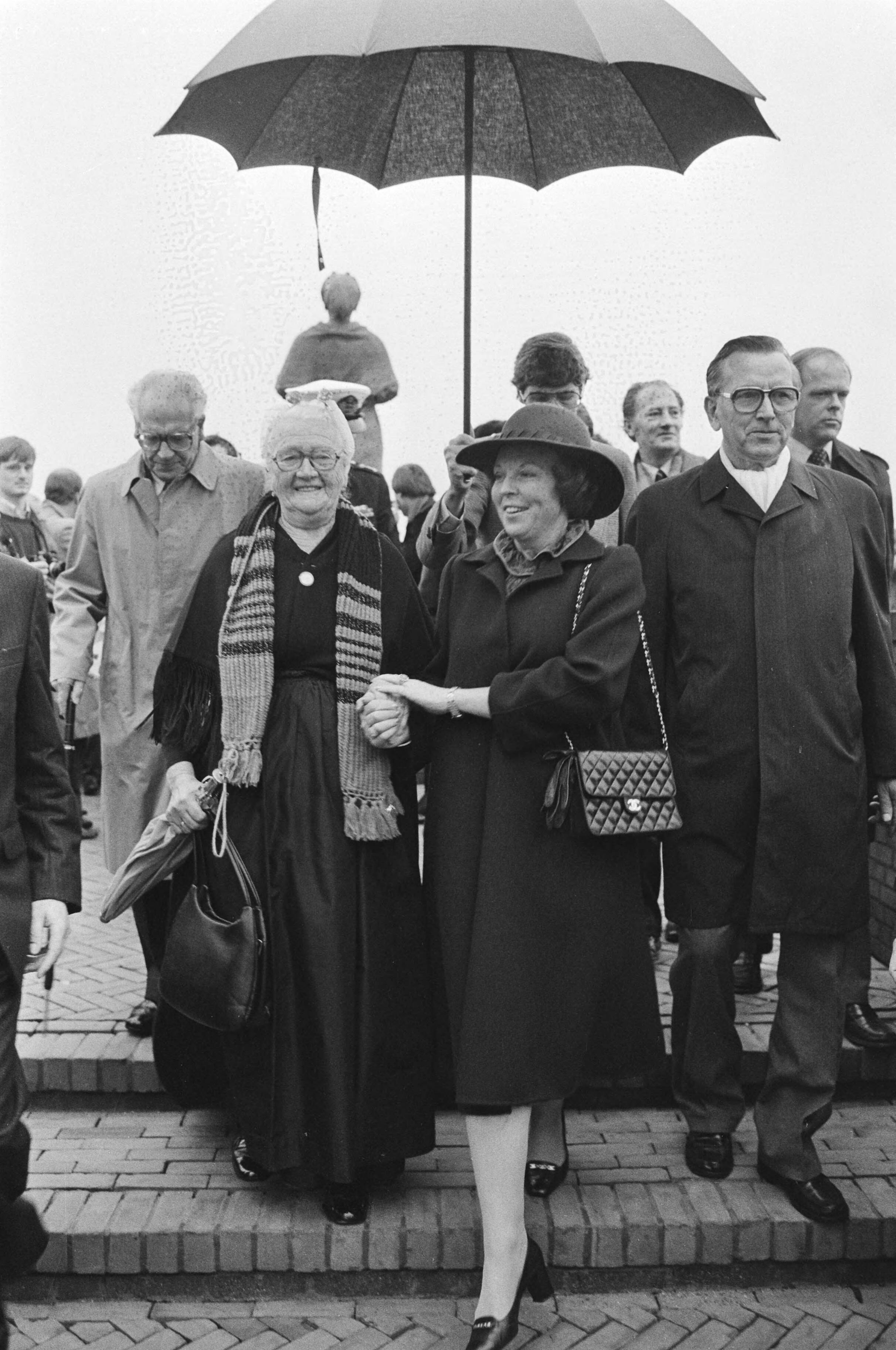 Koningin Beatrix onthult het Vissersmonument in Scheveningen — Koningin Beatrix loopt hand in hand met een Scheveningse vissersvrouw — Foto: Antonisse, Marcel / Anefo. Licentie: Set B: Rechtenvrij / Publiek Domein. Bron: Nationaal Archief. http://hdl.handle.net/10648/ad216bb8-d0b4-102d-bcf8-003048976d84