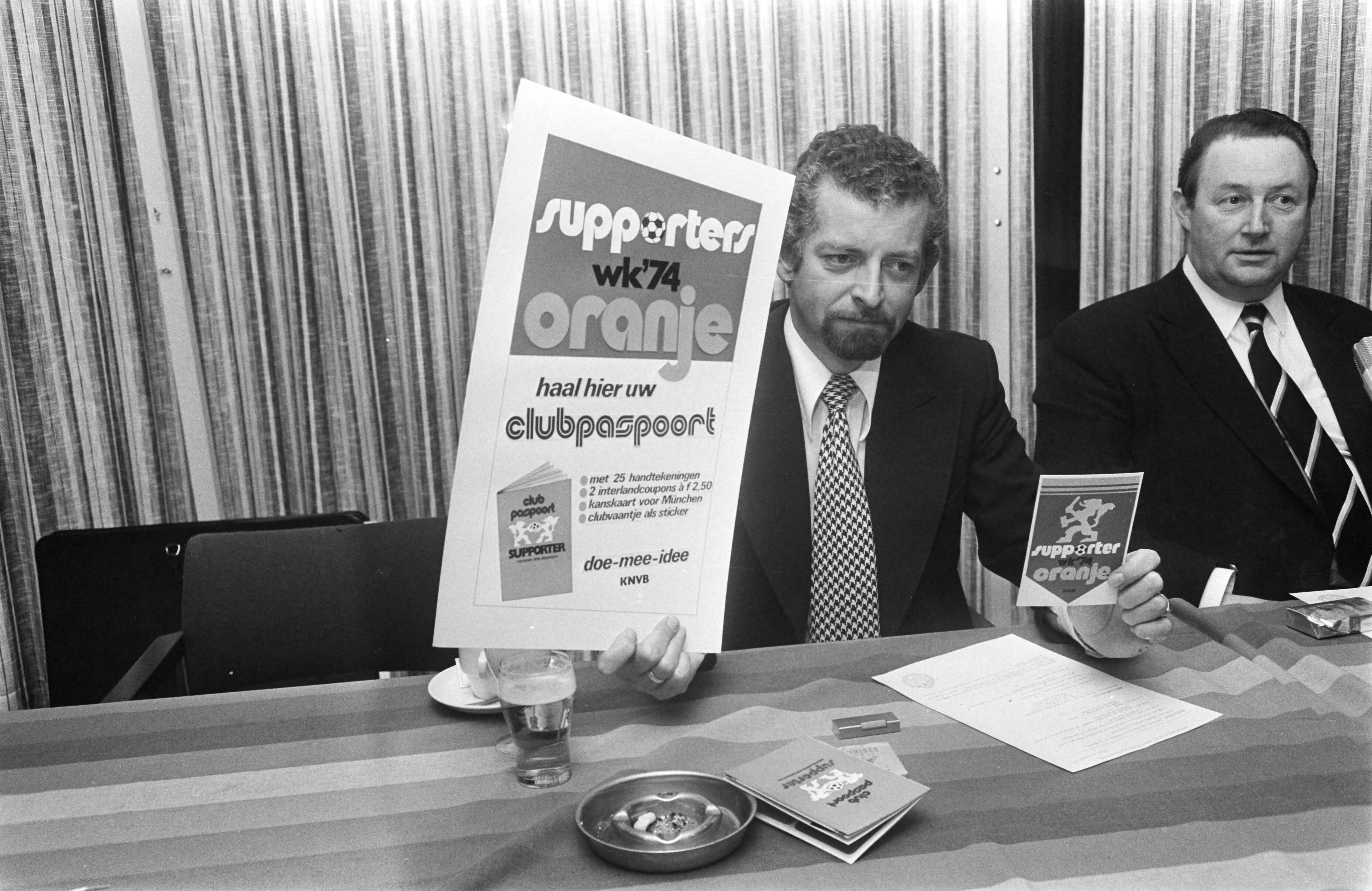 Rinus Michels, supervisor van het Nederlands elftal bij het WK voetbal in 1974 geeft een persconferentie in het KNVB-centrum te Zeist — Marketingdeskundige Maarten van Wamelen met promotieartikelen, rechts van hem KNVB bestuurslid Zwikstra — Foto: Fotograaf Onbekend / Anefo. Licentie: Set B: Rechtenvrij / Publiek Domein. Bron: Nationaal Archief. http://hdl.handle.net/10648/ac42cde0-d0b4-102d-bcf8-003048976d84